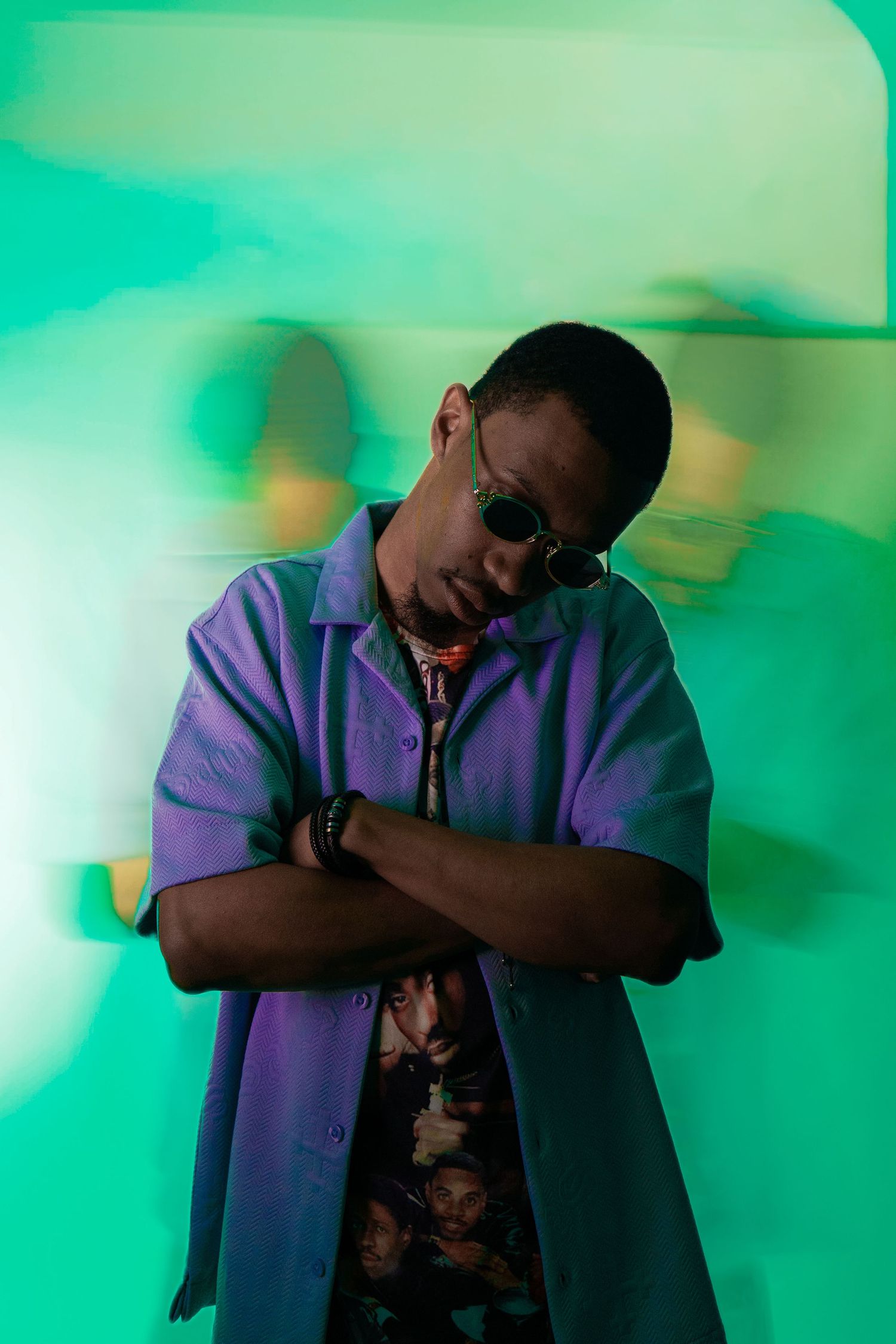 Stylish Black man in purple shirt and sunglasses poses with arms crossed against vibrant green background.