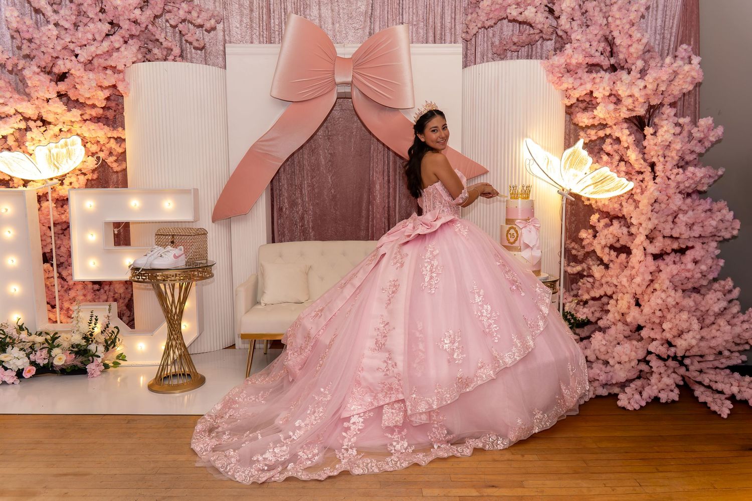 Teen in pink lace ball gown poses before bow backdrop with cherry blossom trees at quinceañera.