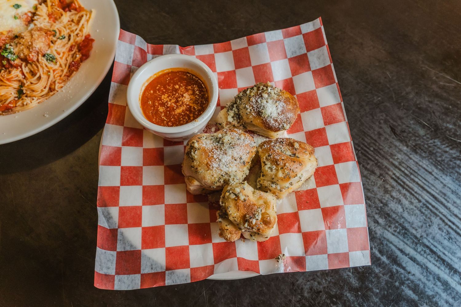 Garlic knots served on red checkered paper with a side of marinara dipping sauce in a basket.