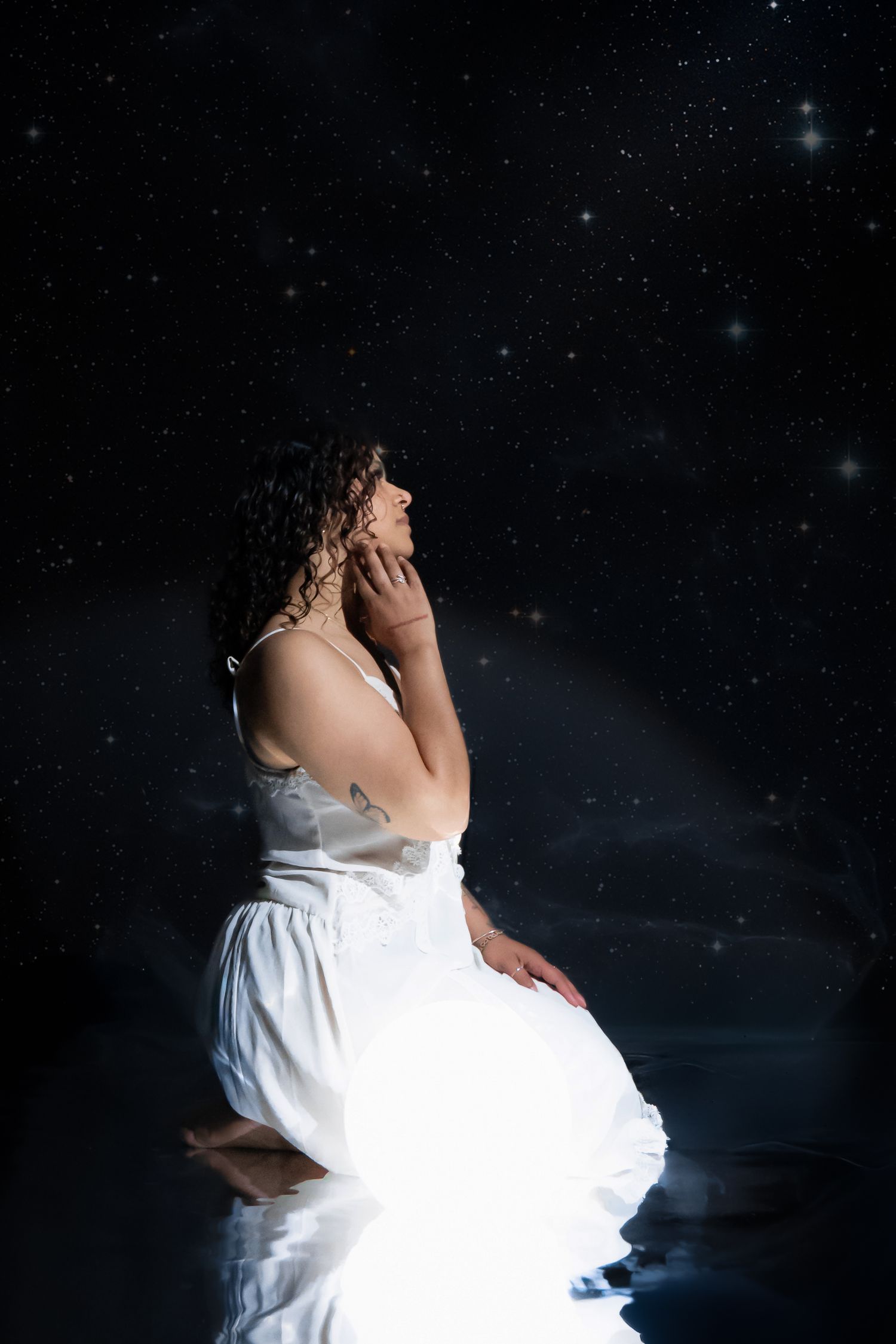 Woman in white dress kneeling under a starry night sky, bathed in soft light, gazing upward in a serene pose.