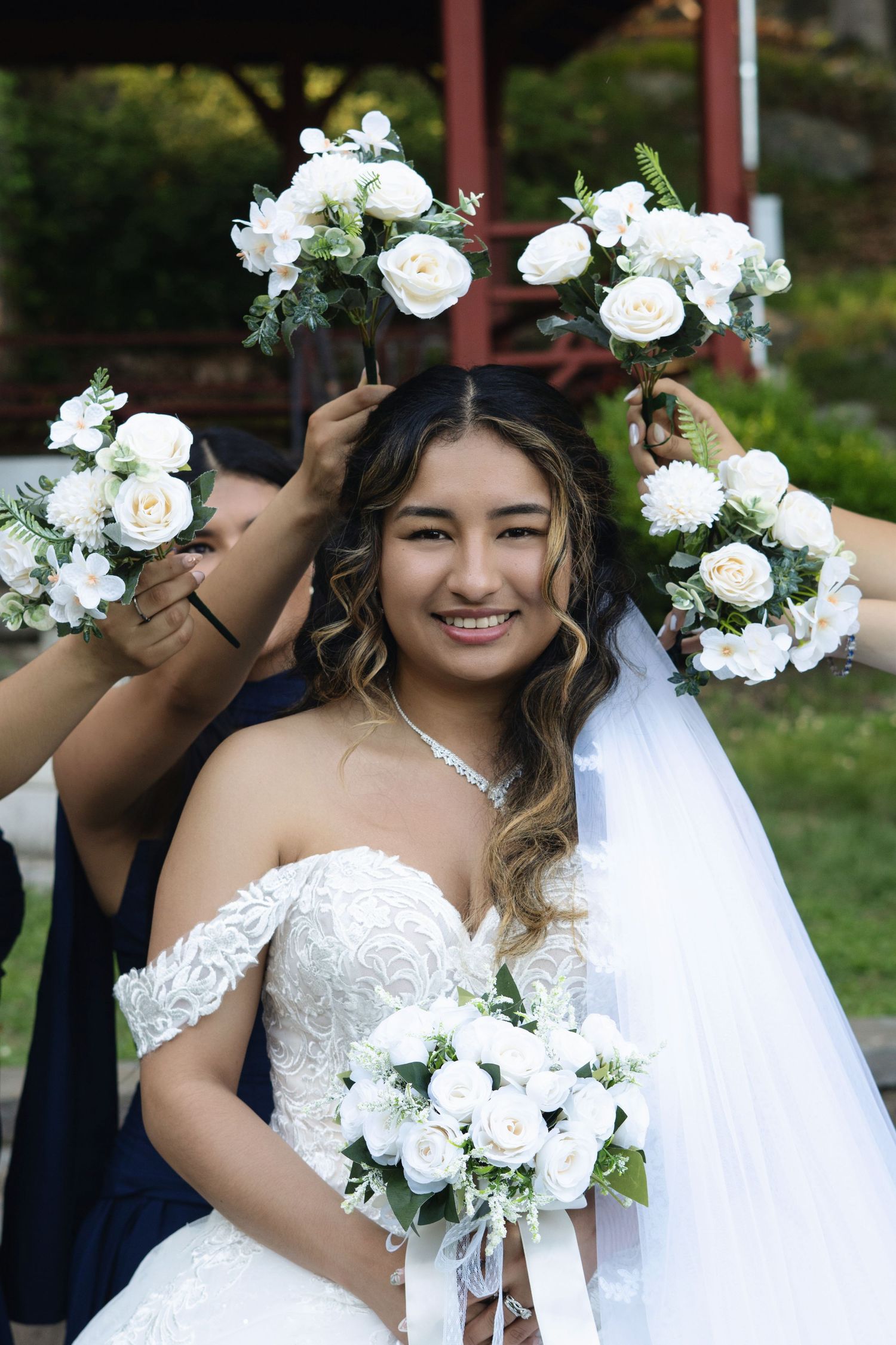 Smiling bride in white lace gown holds bouquet of white roses, surrounded by bridesmaids in navy dresses at outdoor wedding.