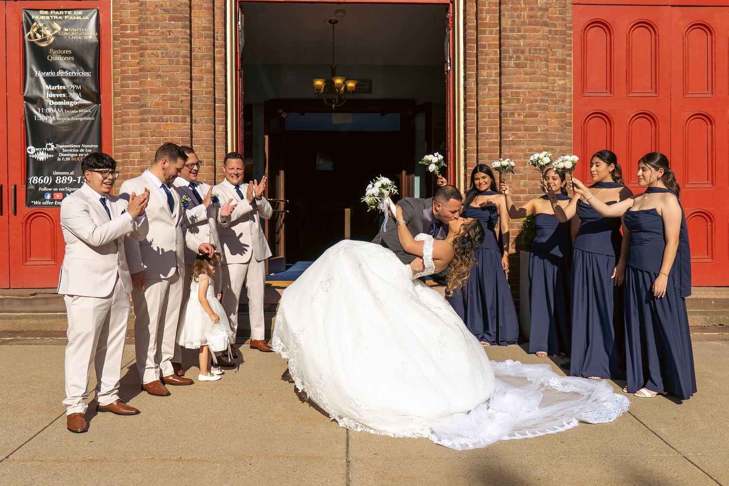 Wedding party poses outside brick church with red doors, bride in white gown, bridesmaids in navy blue dresses, groomsmen in tan suits.