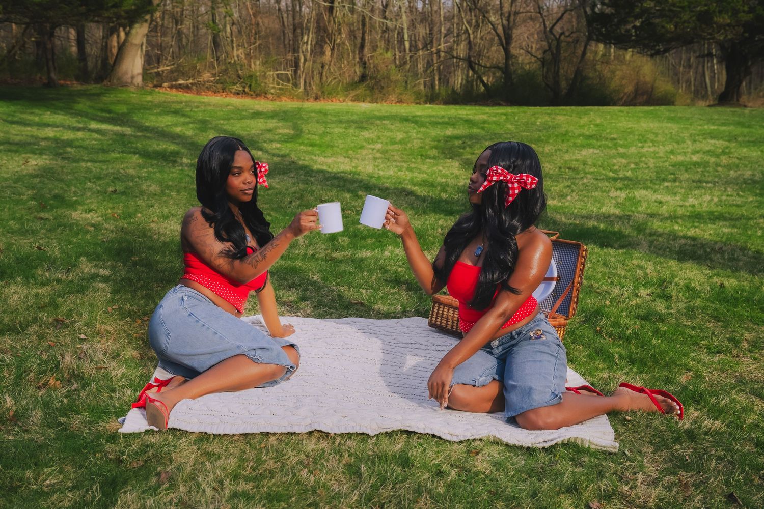 Two women in matching red outfits enjoy a picnic on a blanket in a park, toasting with mugs.