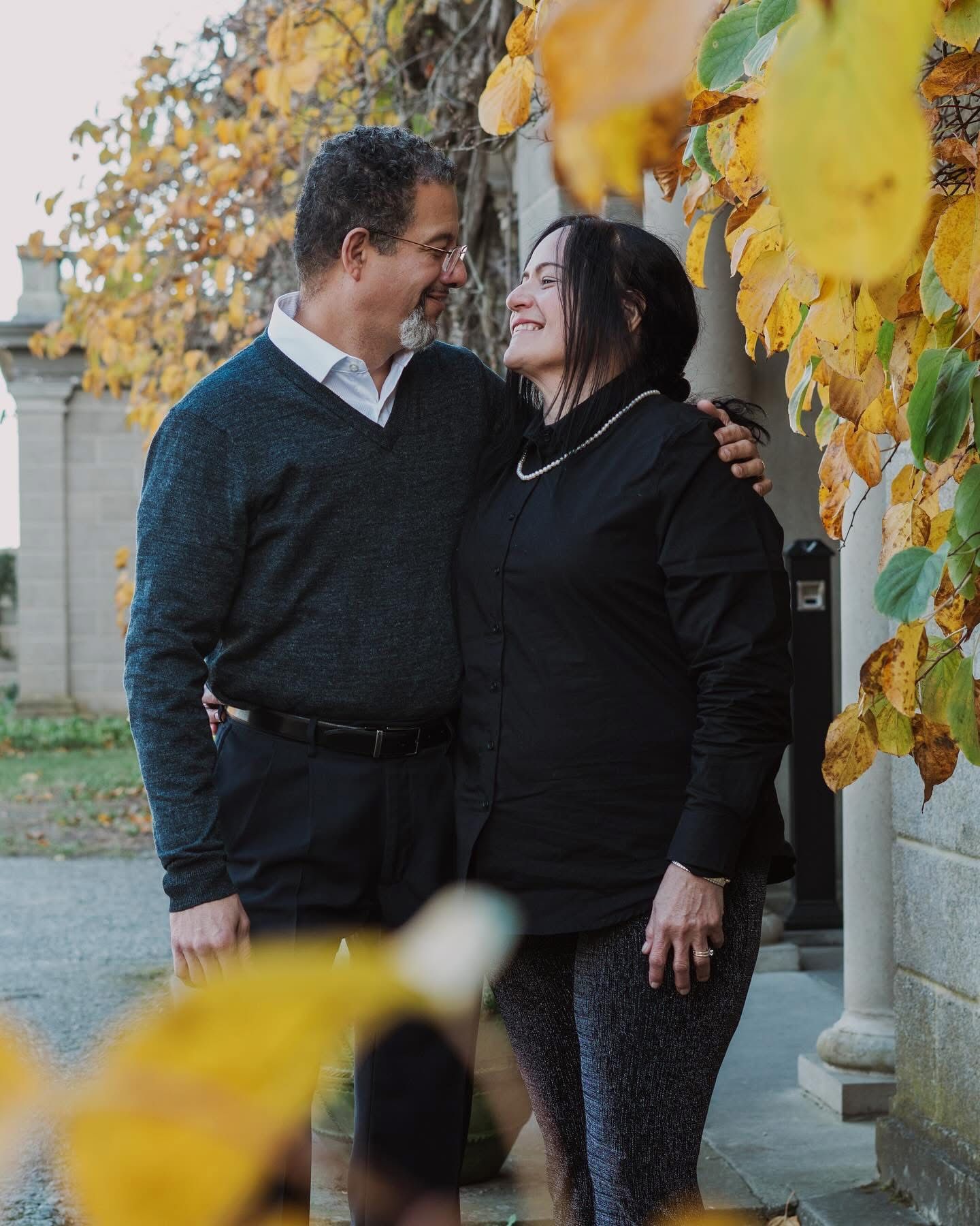 Happy couple smiling at each other surrounded by golden autumn leaves outside a stone building.