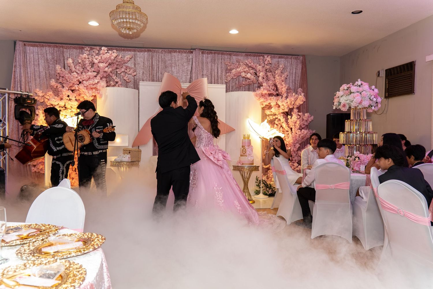 Mariachi band plays as quinceañera girl in pink ball gown dances on fog-filled floor at reception.