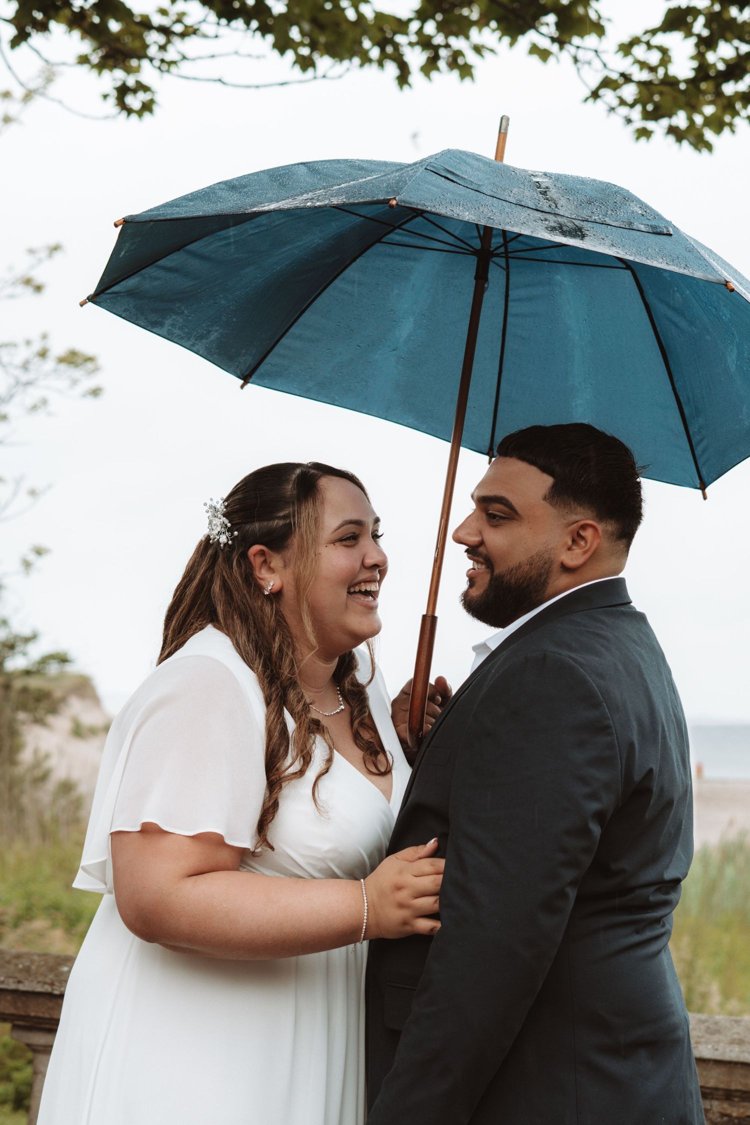 Happy bride and groom smiling at each other under a blue umbrella during their rainy outdoor wedding.