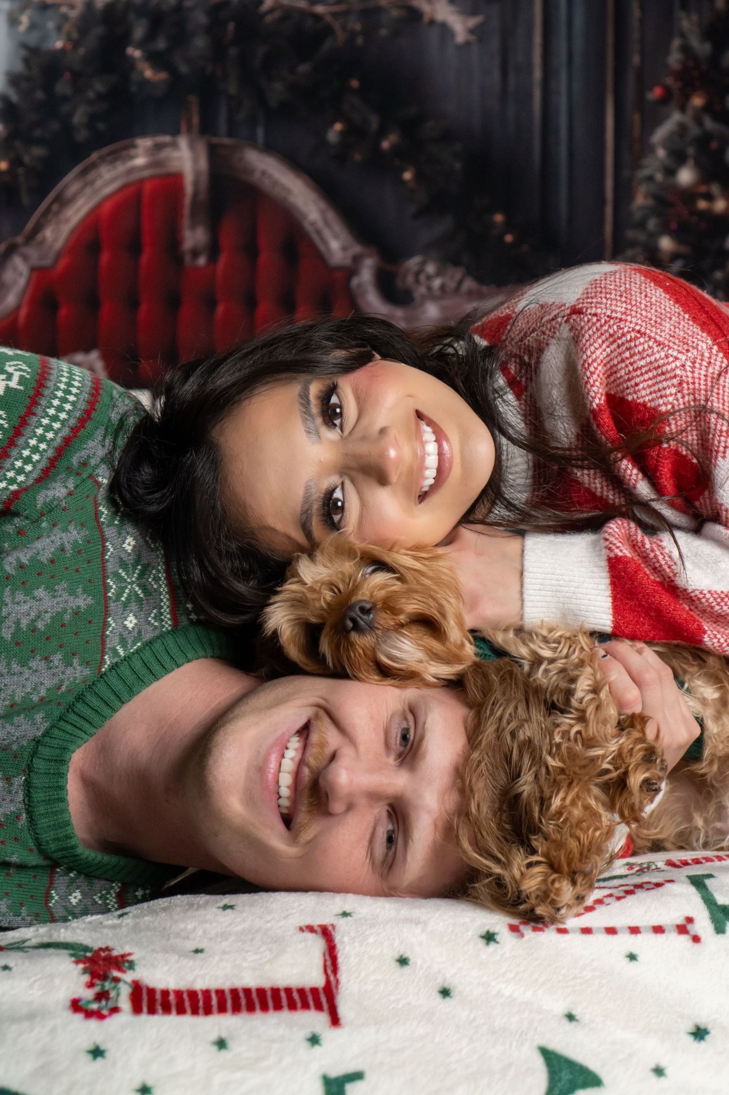 Smiling couple in Christmas sweaters lying with small brown dog near holiday decorations.