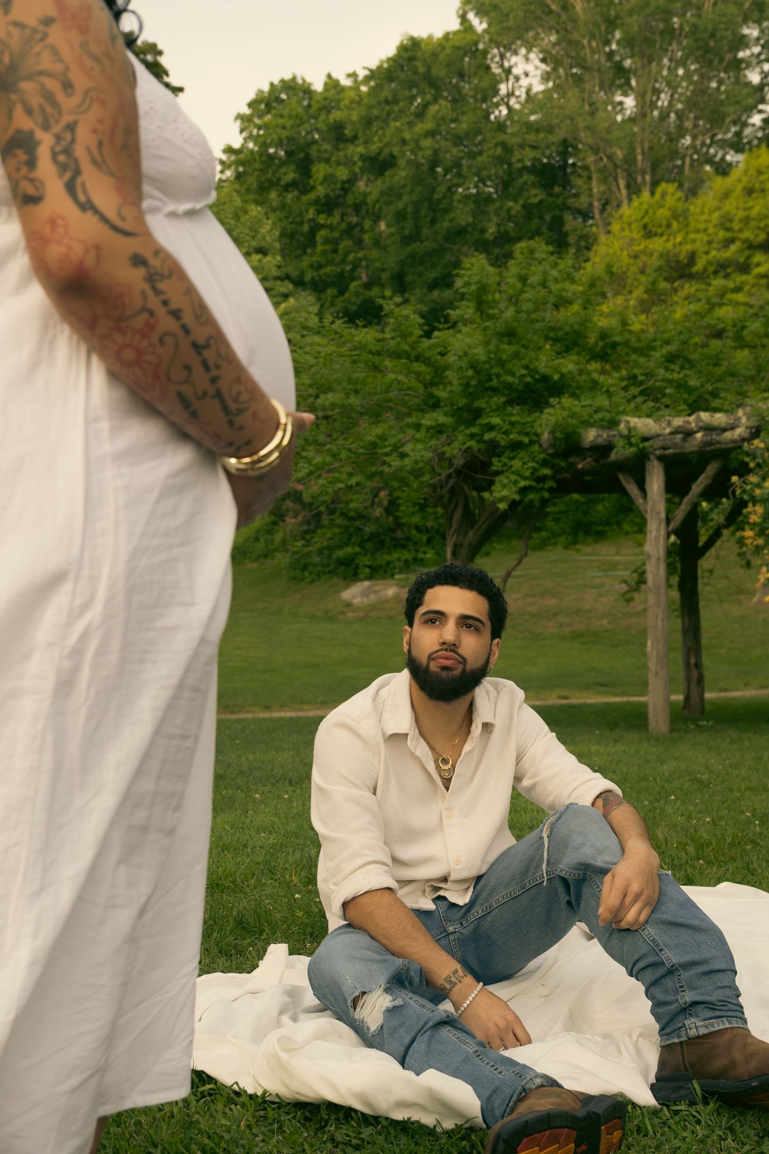 A man sits on a blanket in a park, looking up at a tattooed pregnant woman in a white dress.