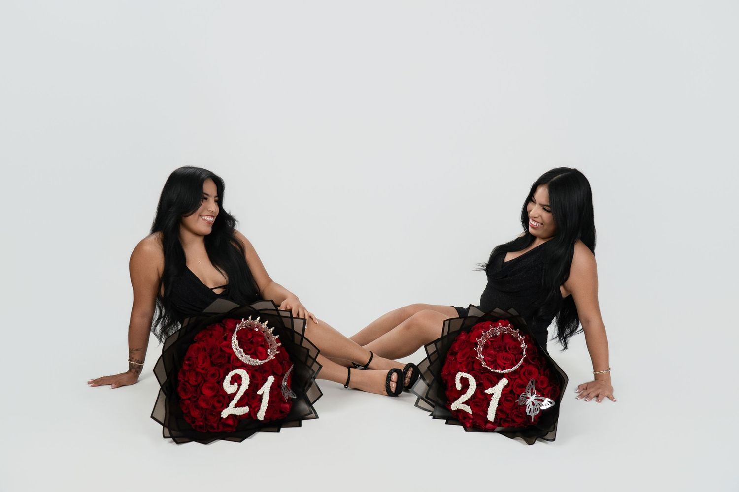 Two women in black dresses sit with red rose bouquets numbered 21, celebrating a 21st birthday against a white background.