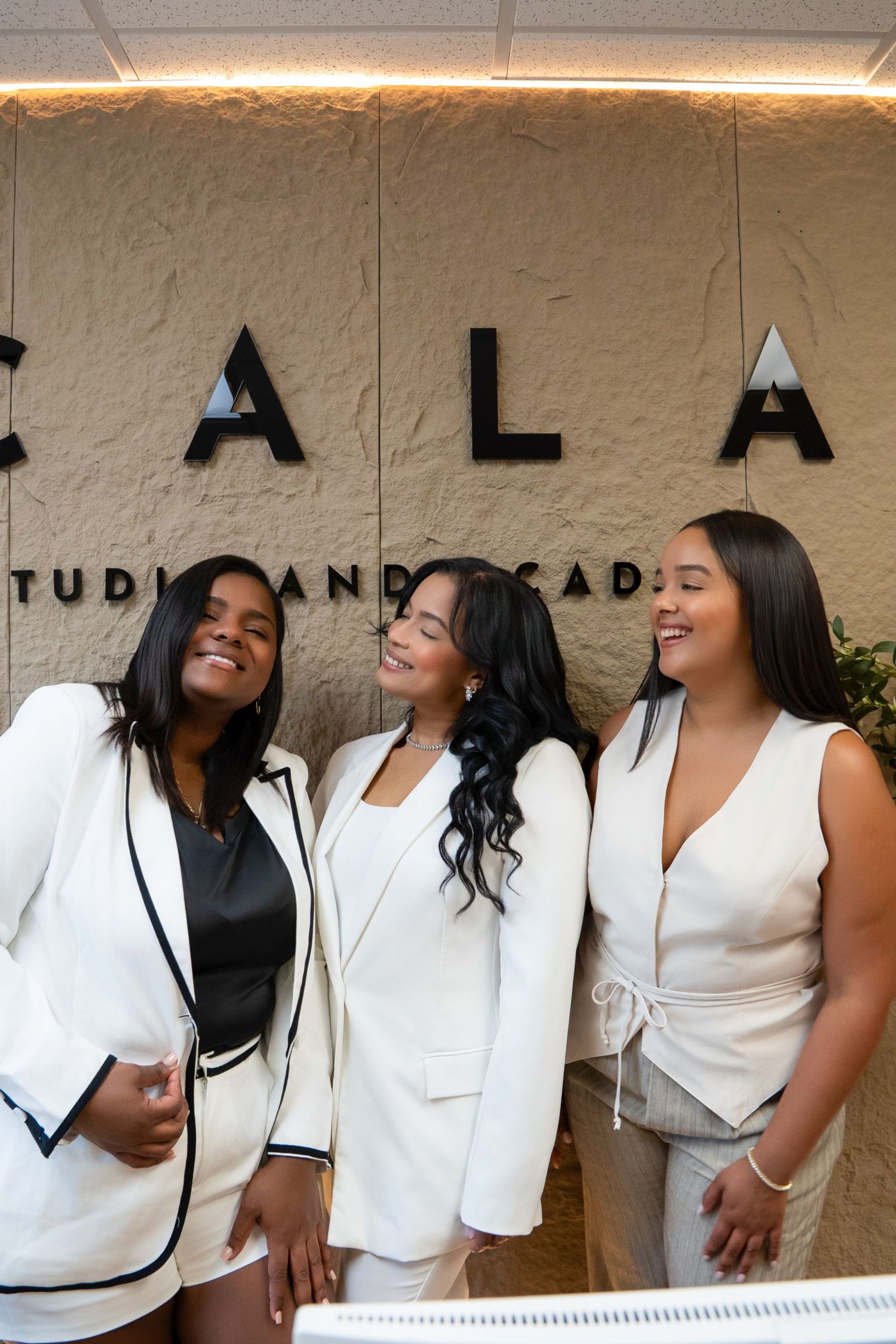 Three women in white blazers smile together in front of a studio academy sign.