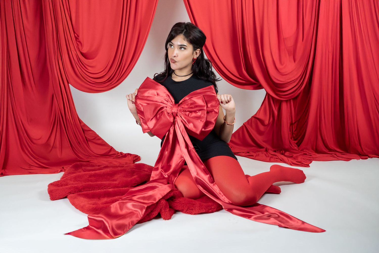 Woman in black bodysuit sitting with large red satin bow against red drape backdrop.