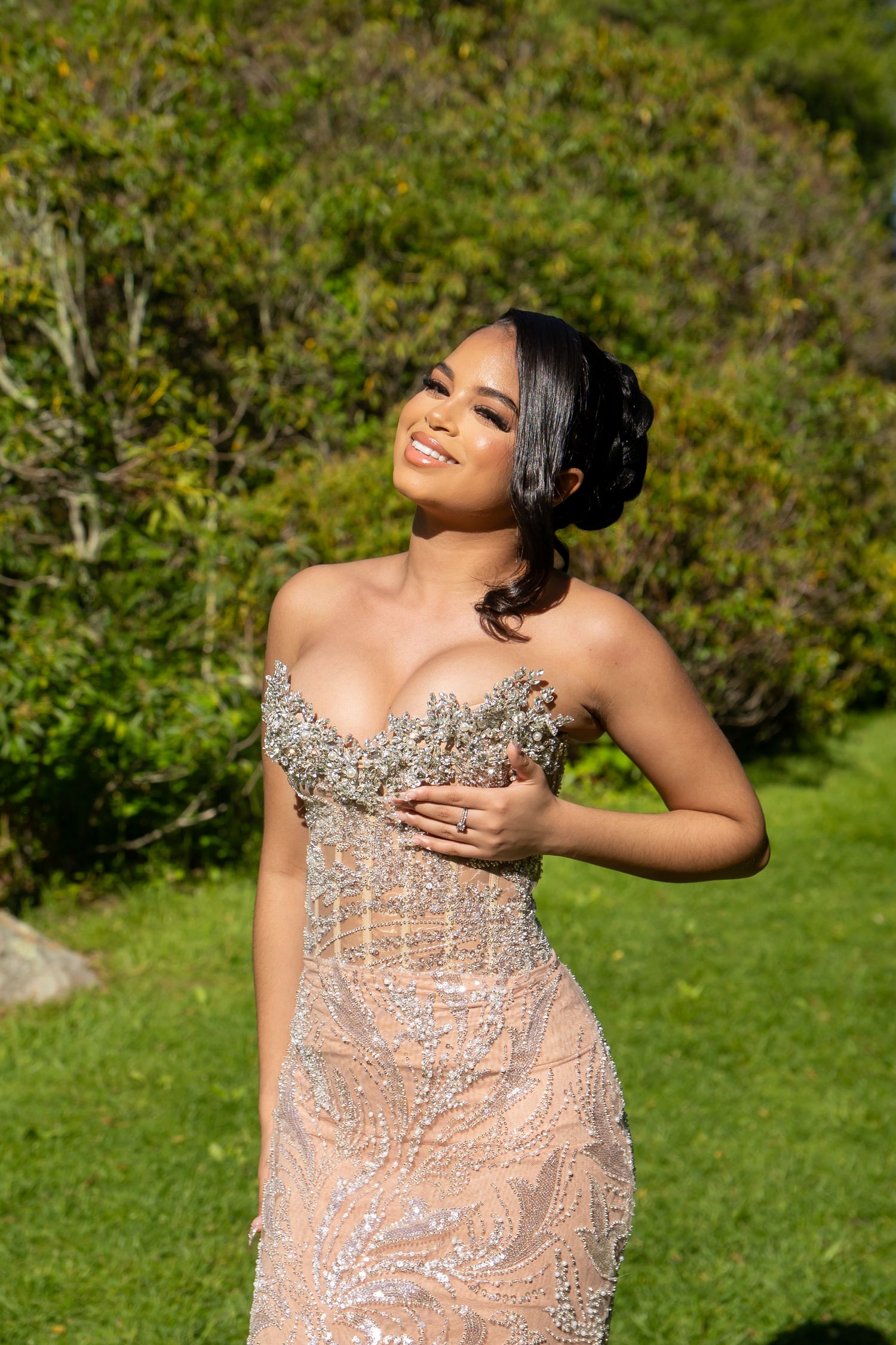Young woman in a beaded strapless gown smiling outdoors in a lush green garden setting.