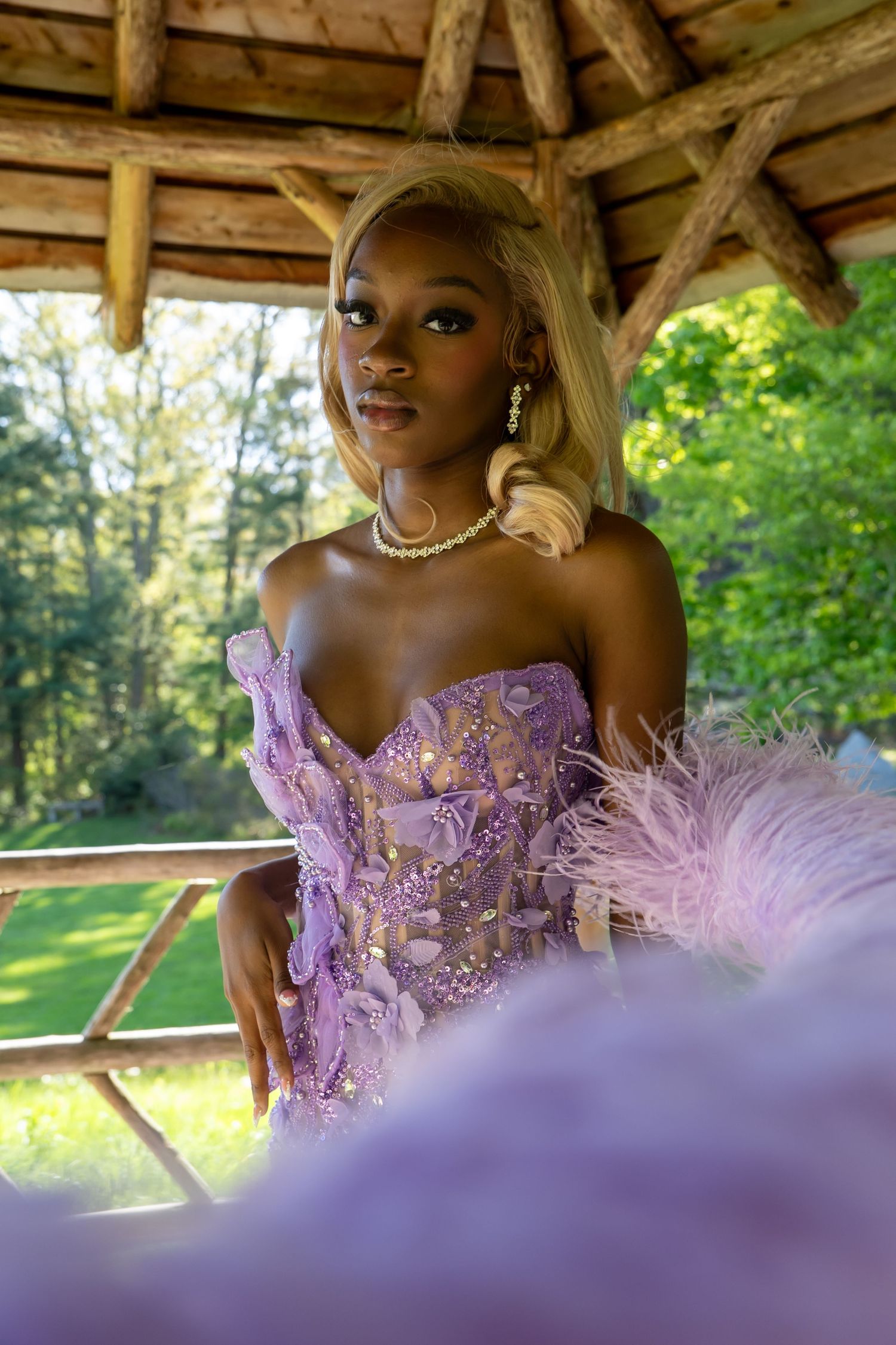 Young woman in lavender feathered gown with pearl necklace posing under rustic gazebo surrounded by lush greenery.