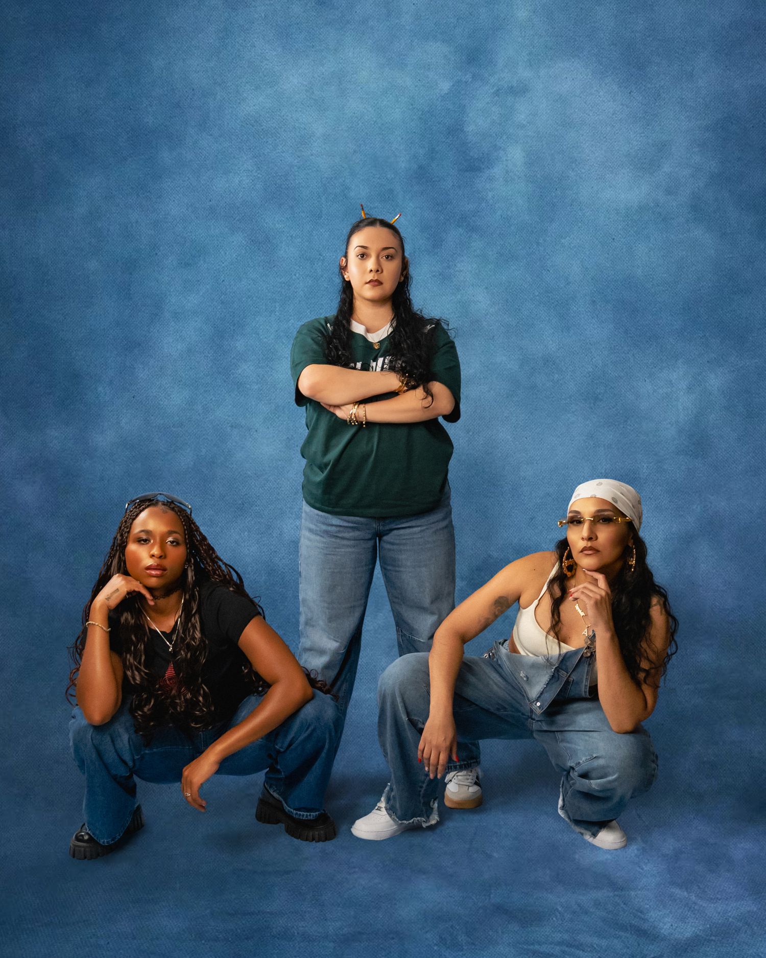 Three young women in jeans pose against a blue mottled studio backdrop, one standing with arms crossed and two crouching on either side.