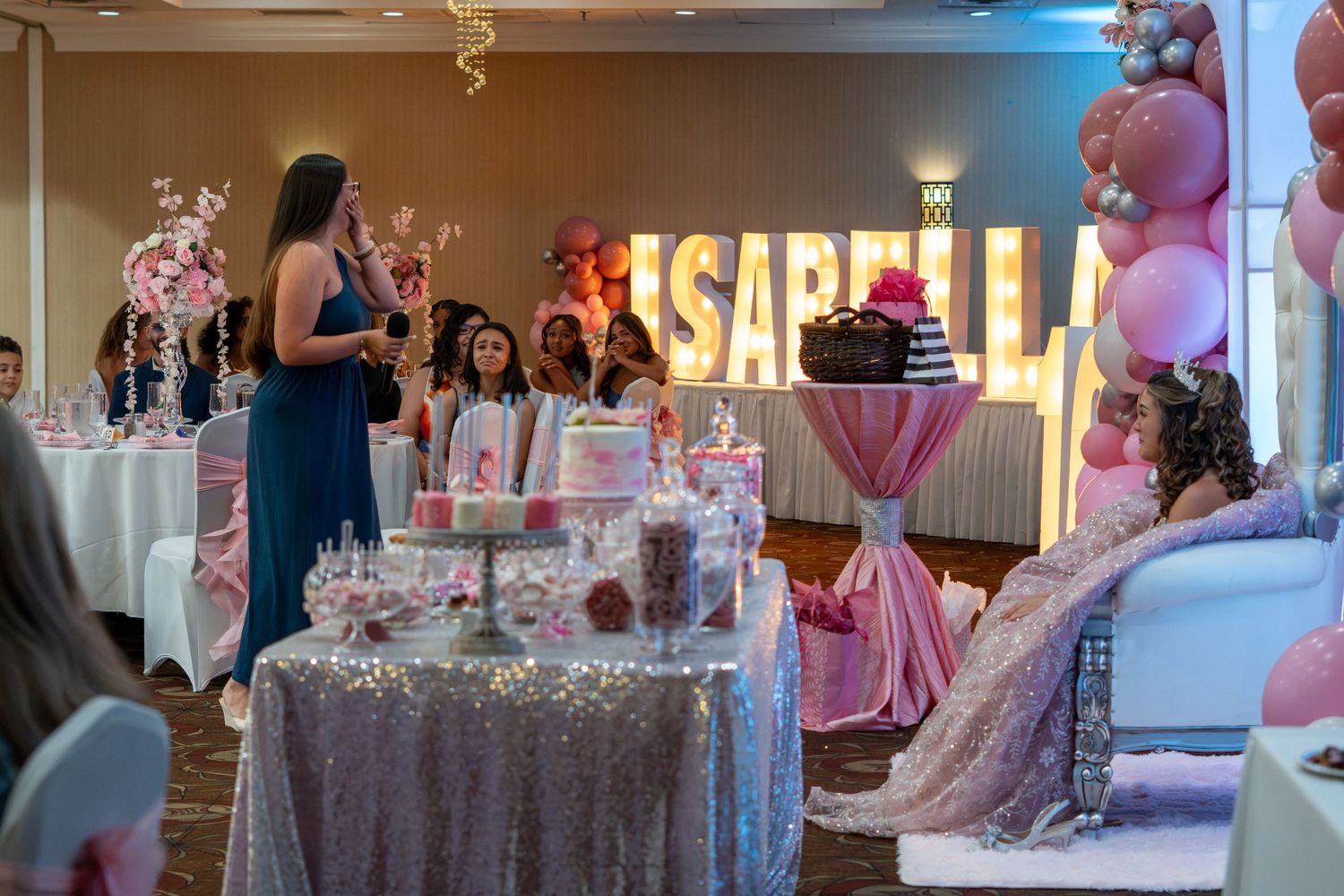 A woman in blue dress presents an elegant pink dessert table at Isabella's quinceañera celebration. Sweet 16!