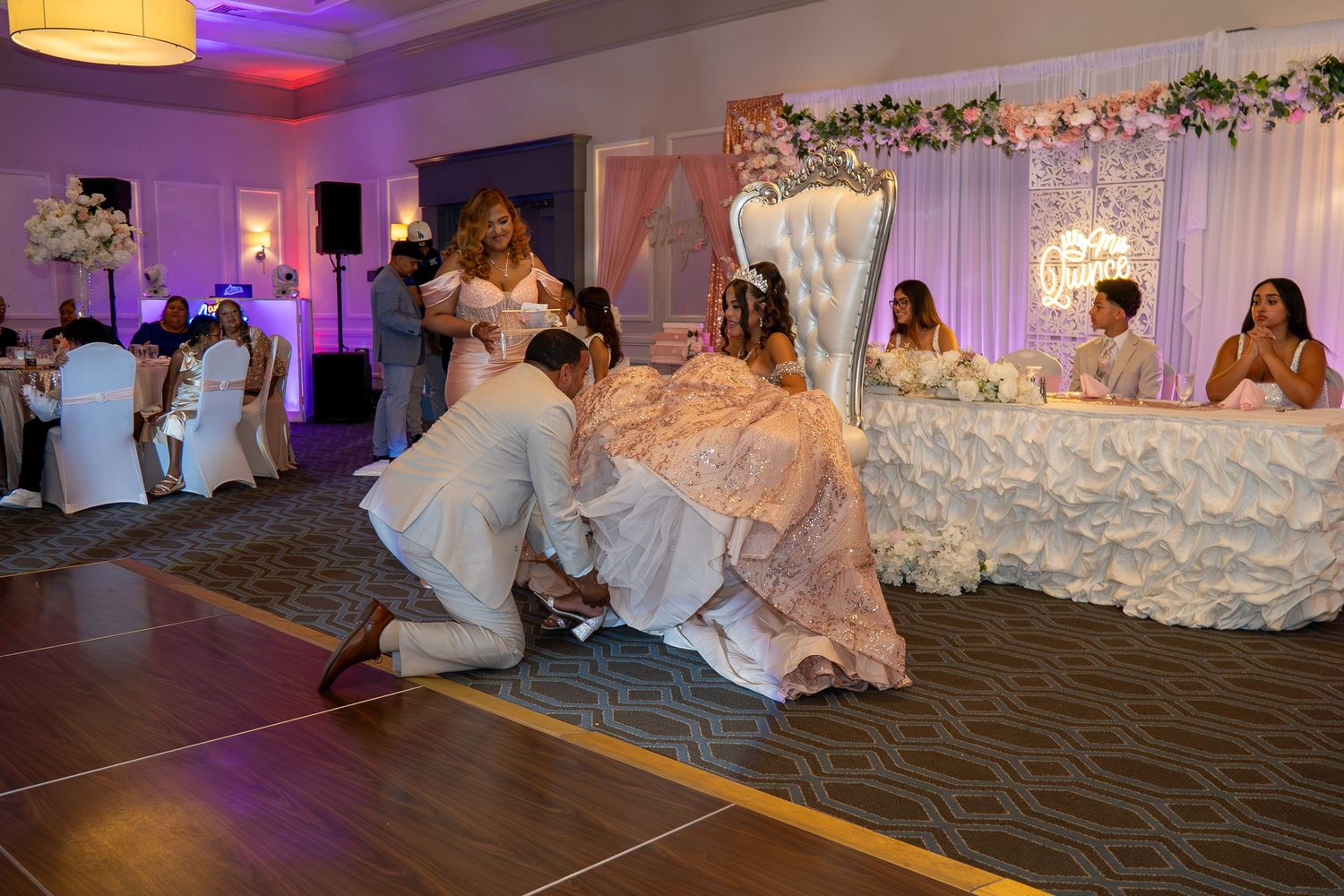 A groom kneels before his bride in a pink gown during a wedding reception in an elegantly decorated ballroom. Sweet 16/Quinceañera
