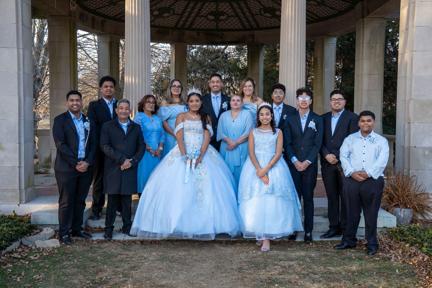 Group photo of families celebrating a quinceañera, with girls in blue ball gowns posing under a stone gazebo outdoors. Harkness Park Sweet15