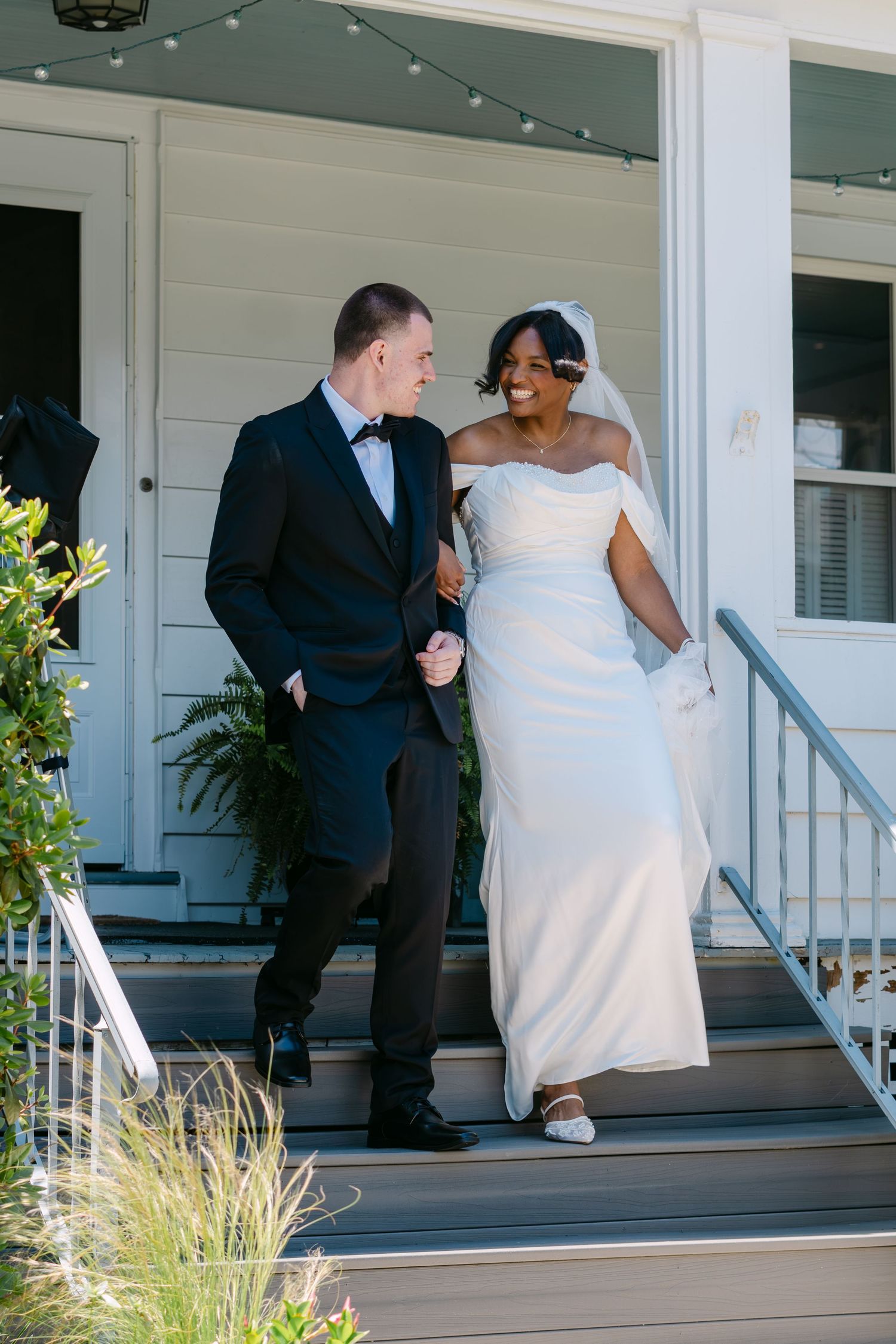 Bride and groom walking down porch steps, smiling at each other, she in white off-shoulder gown, he in black tuxedo.