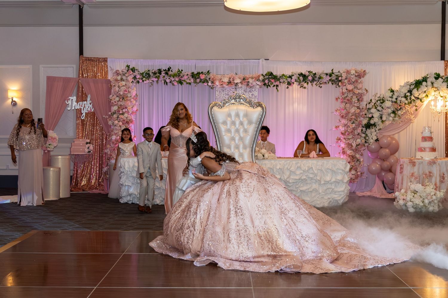 Quinceañera girl in pink ballgown poses on throne stage with floral backdrop and guests. Sweet 16/Quinceañera