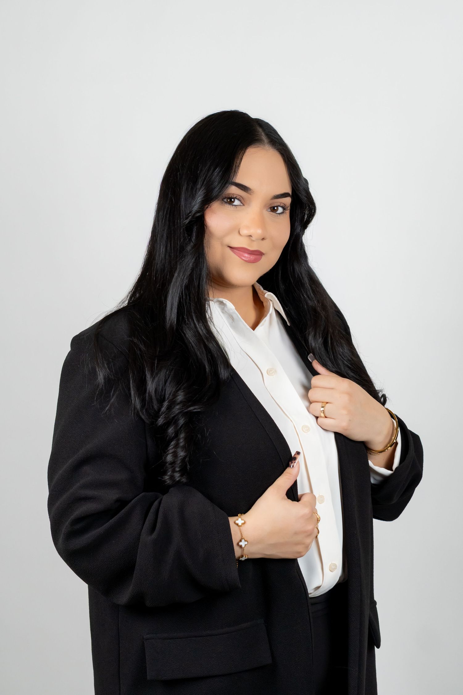 Professional woman in black blazer and white shirt poses confidently against light gray background.