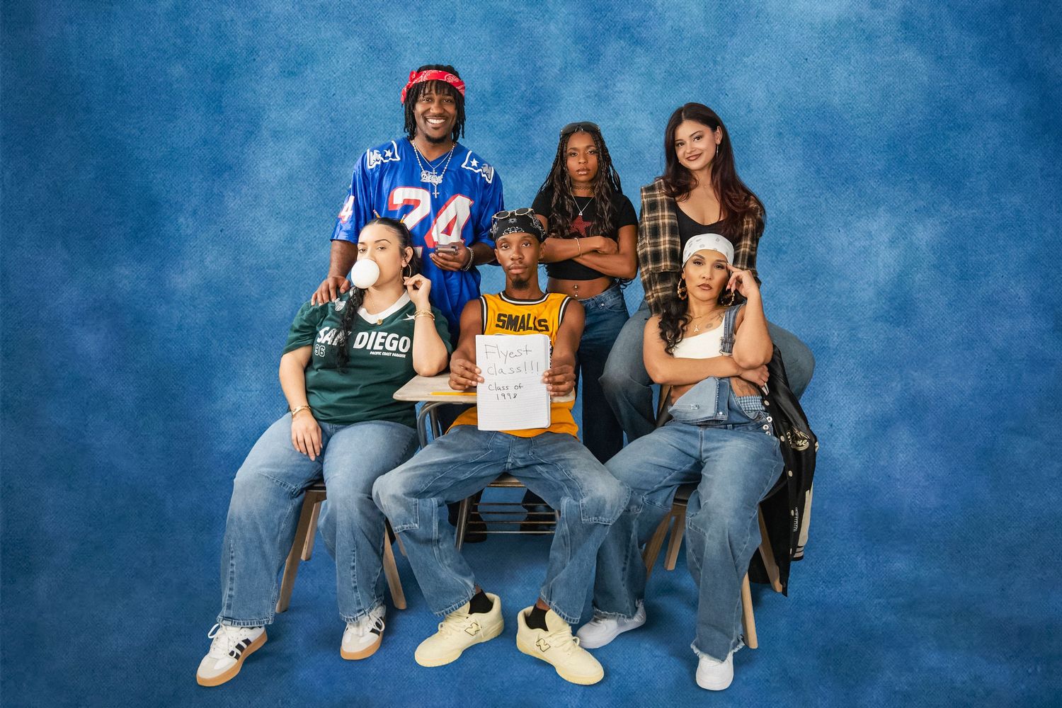 Five young people in casual streetwear, including jerseys and denim, pose together on stools against a textured blue studio backdrop.