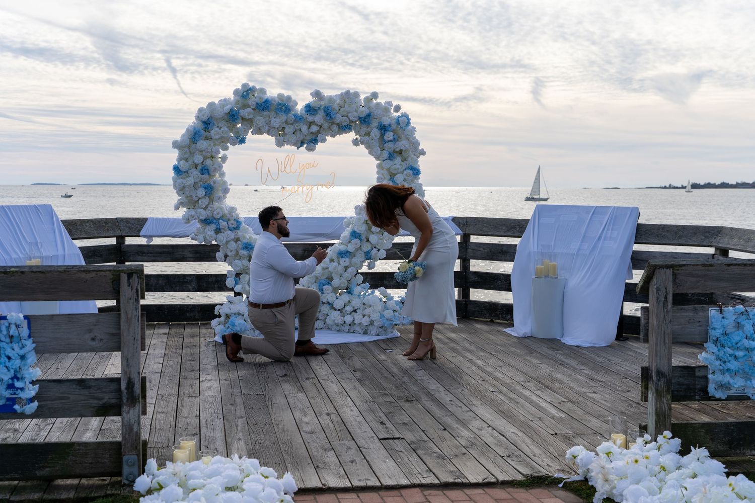 Man kneeling in a marriage proposal to a woman beneath a white and blue floral heart arch on a seaside wooden pier at sunset.