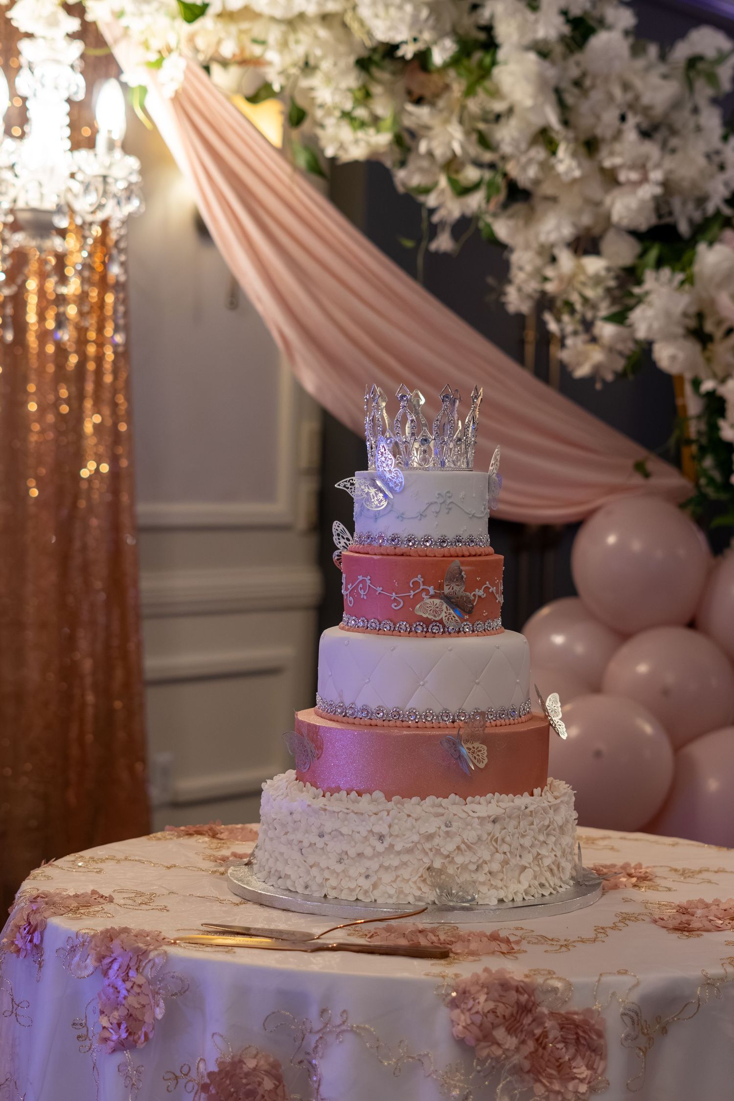 Four-tier pink and white wedding cake with crystal crown and butterfly decorations on floral tablecloth. Sweet 16/Quinceañera