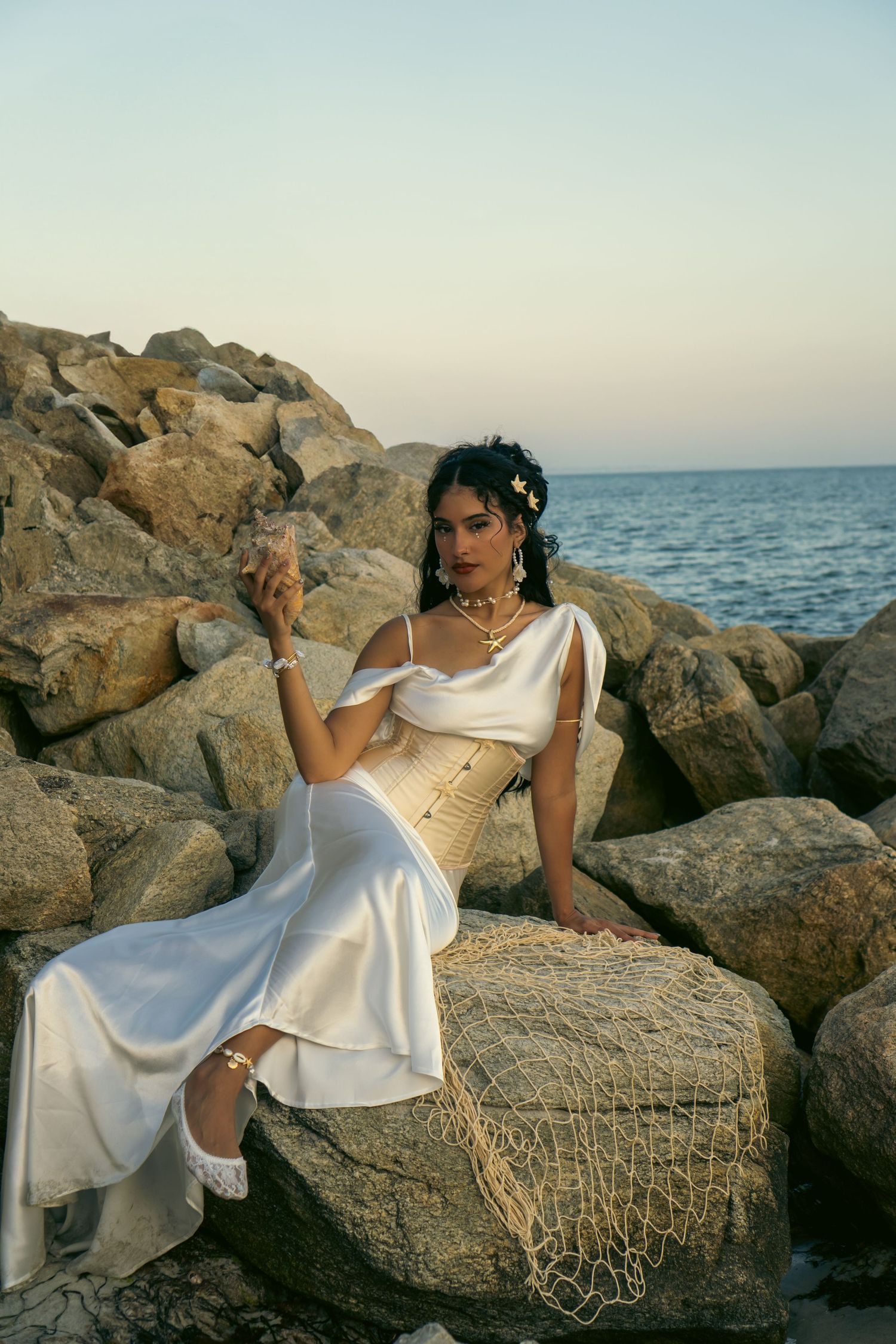 Woman in white satin dress and gold corset poses on coastal rocks by the sea at sunset.
