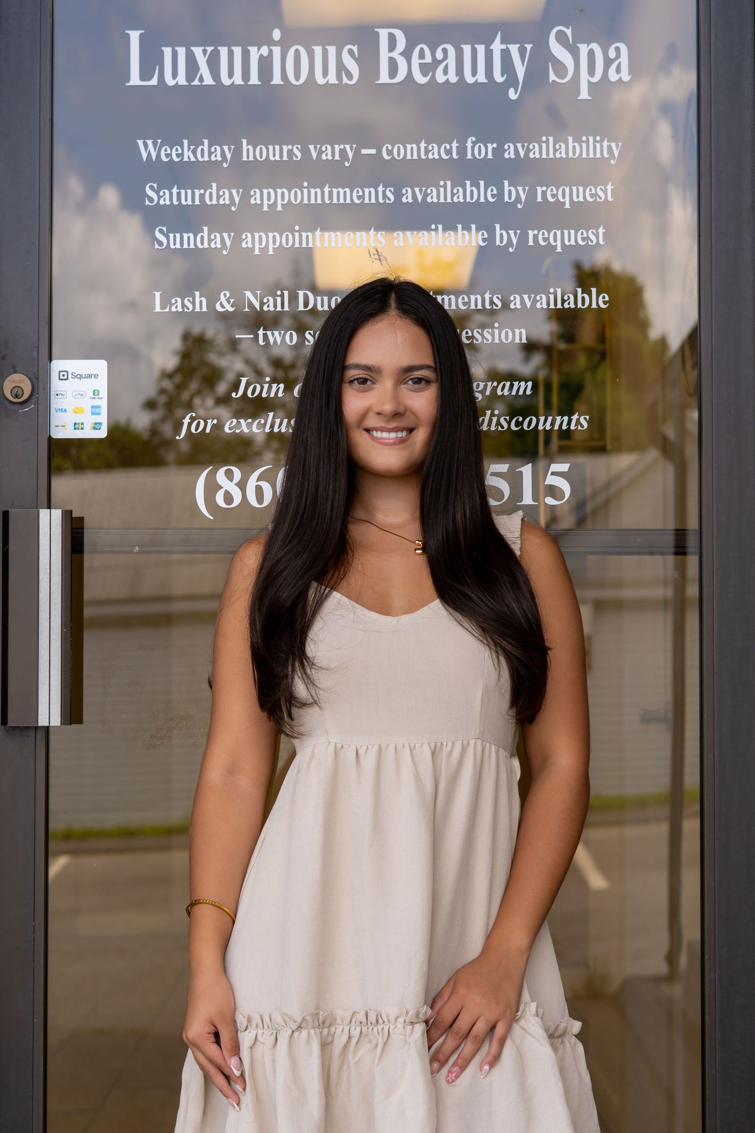 Smiling young woman in white dress standing outside Luxurious Beauty Spa entrance door.
