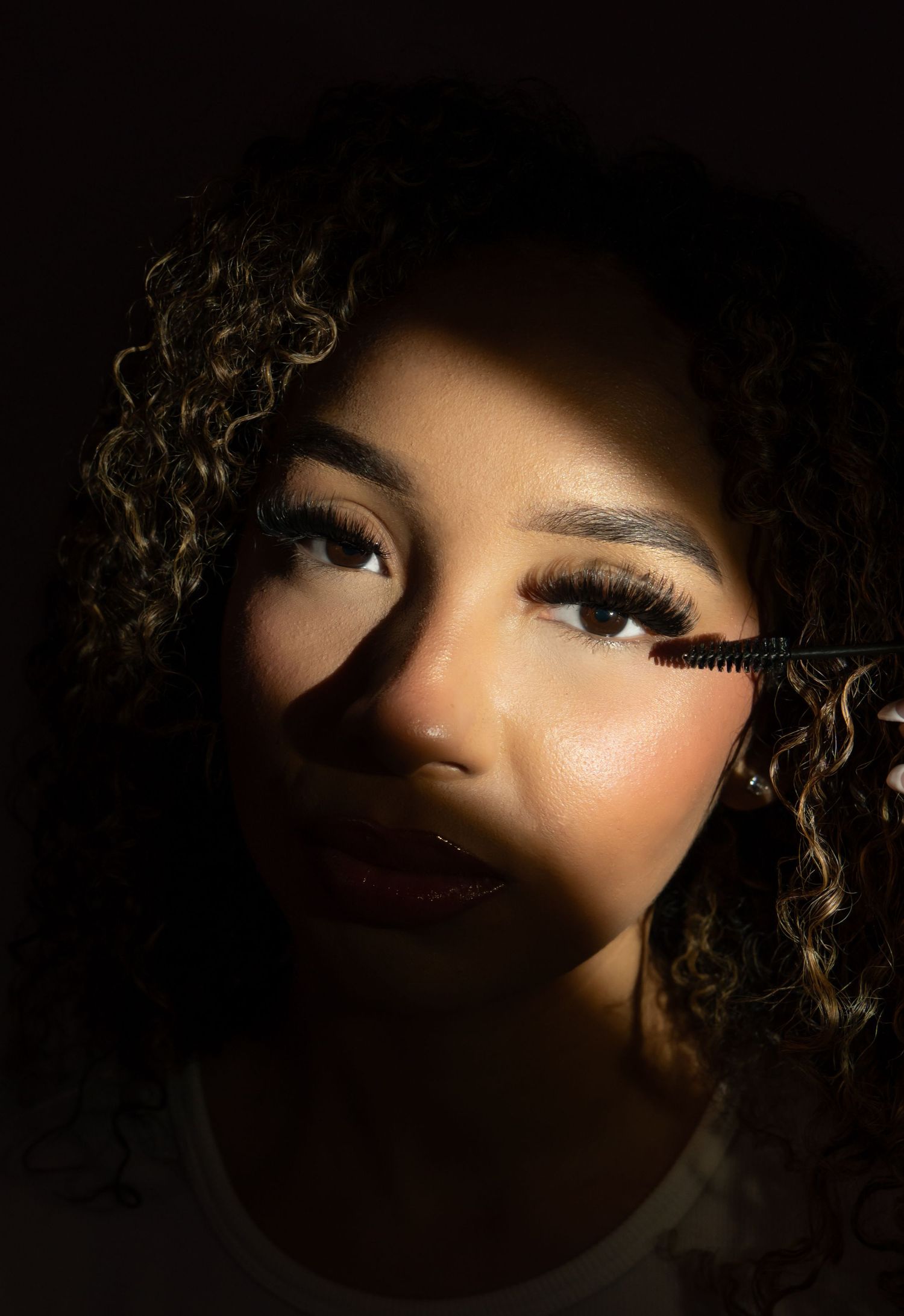 Young woman with curly hair showcases bold lash extensions with mascara wand in dramatic lighting.