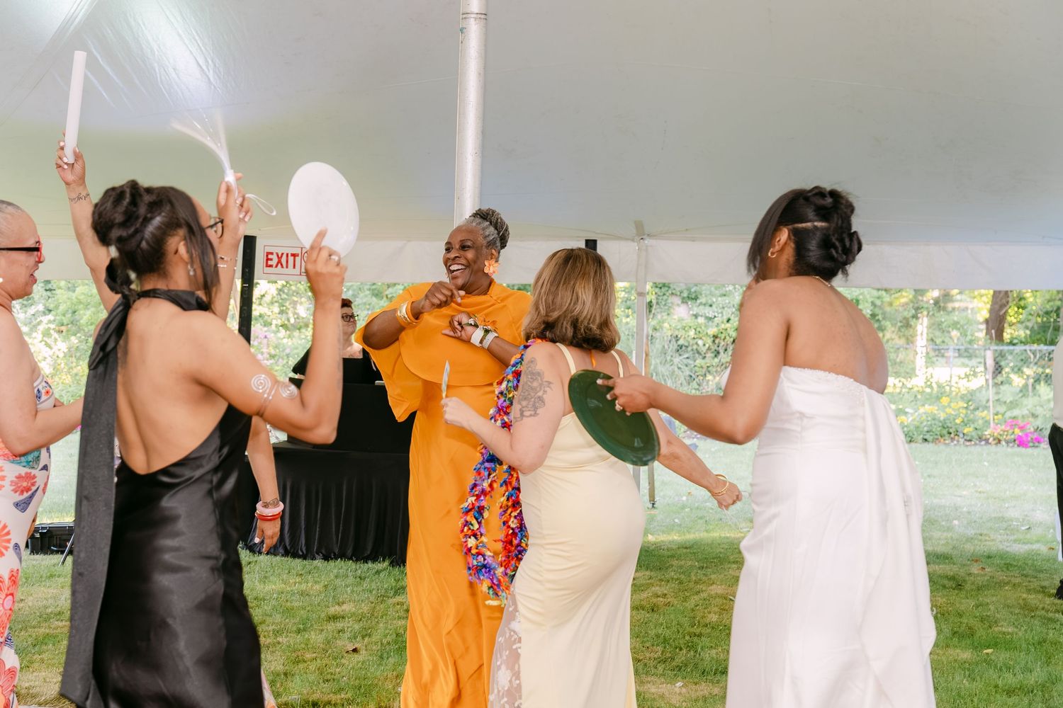 Women dancing joyfully at an outdoor wedding reception under a white tent, wearing elegant dresses.