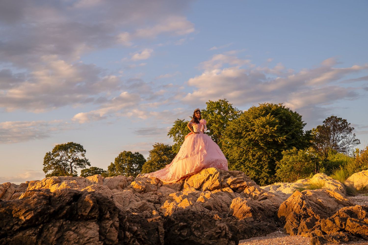 Woman in pink ball gown poses on rocks at golden hour with trees behind.