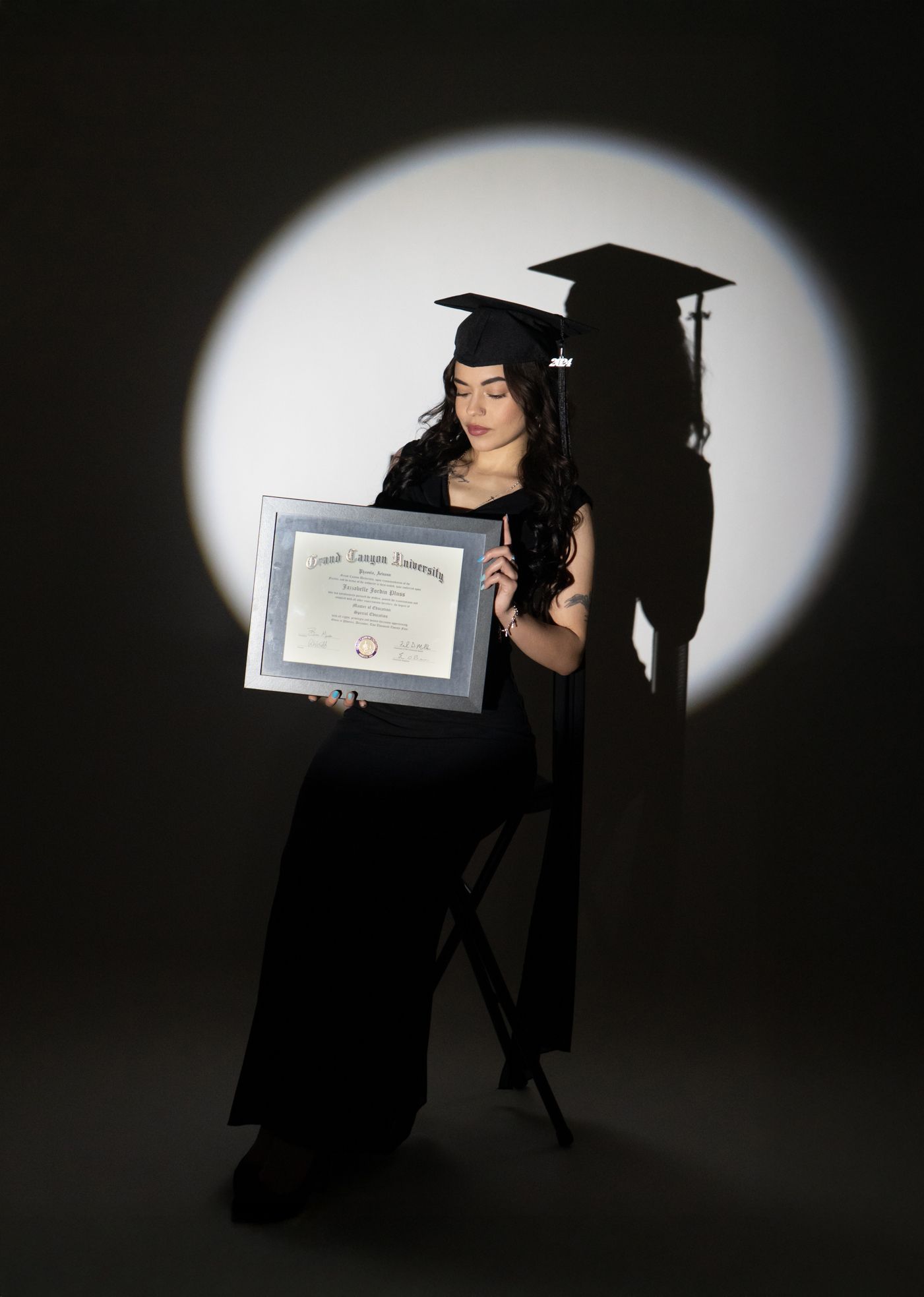 Woman in graduation cap and gown holds framed diploma under spotlight with dramatic shadow backdrop.