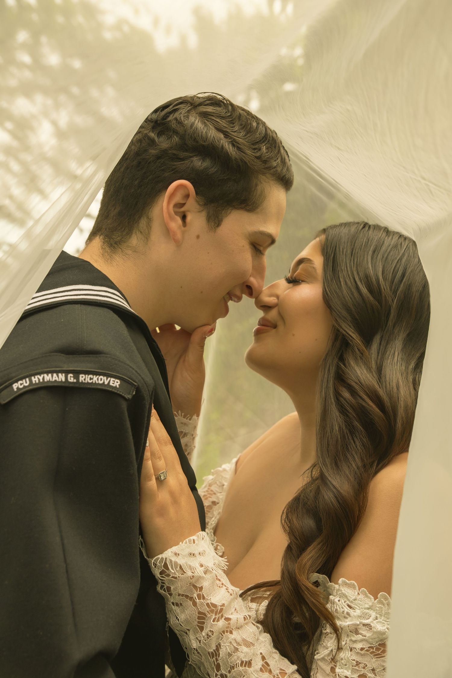 A Navy sailor in uniform nearly kisses his bride in a white lace gown beneath a flowing veil outdoors.
