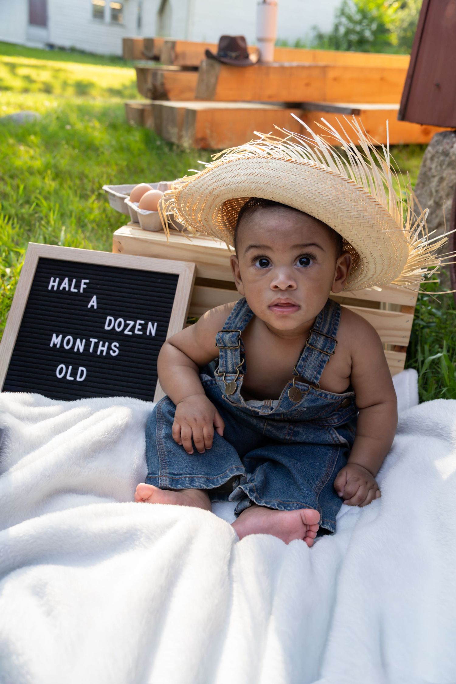 Adorable baby in denim overalls and straw hat sits beside a 'Half A Dozen Months Old' sign on a white blanket outdoors. 6 month photoshoot