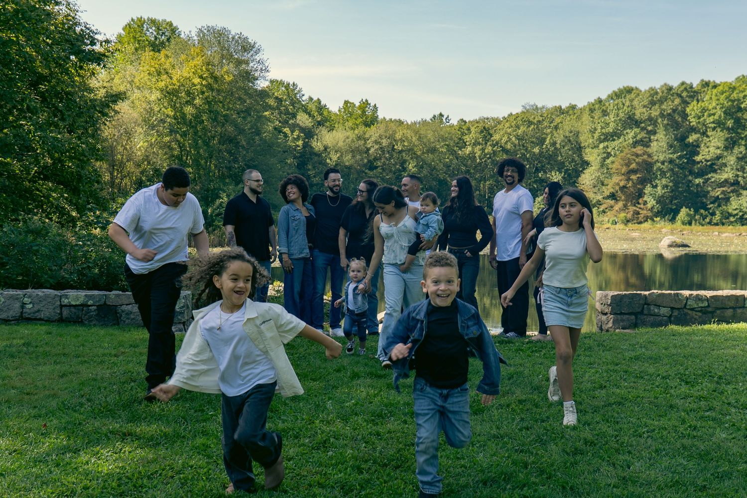 Children run joyfully across a green lawn toward the camera while a group of adults walks behind them near a calm pond surrounded by trees.