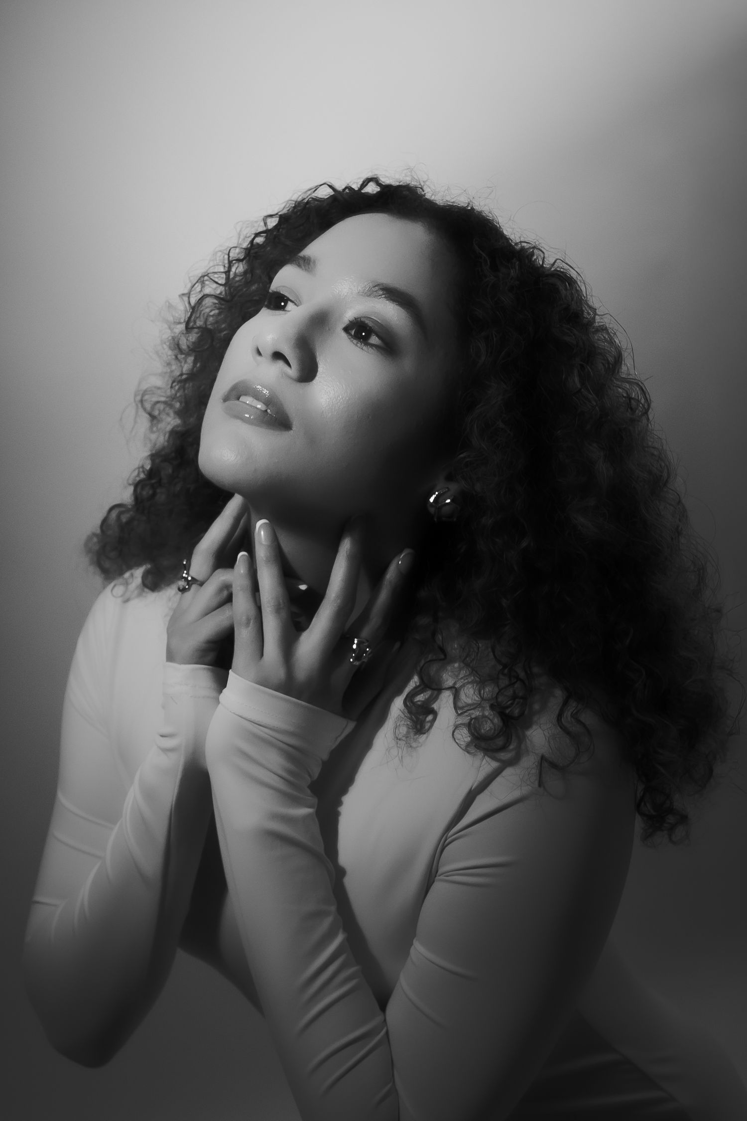 Black and white portrait of a young woman with curly hair, hands gracefully touching her neck, gazing upward.