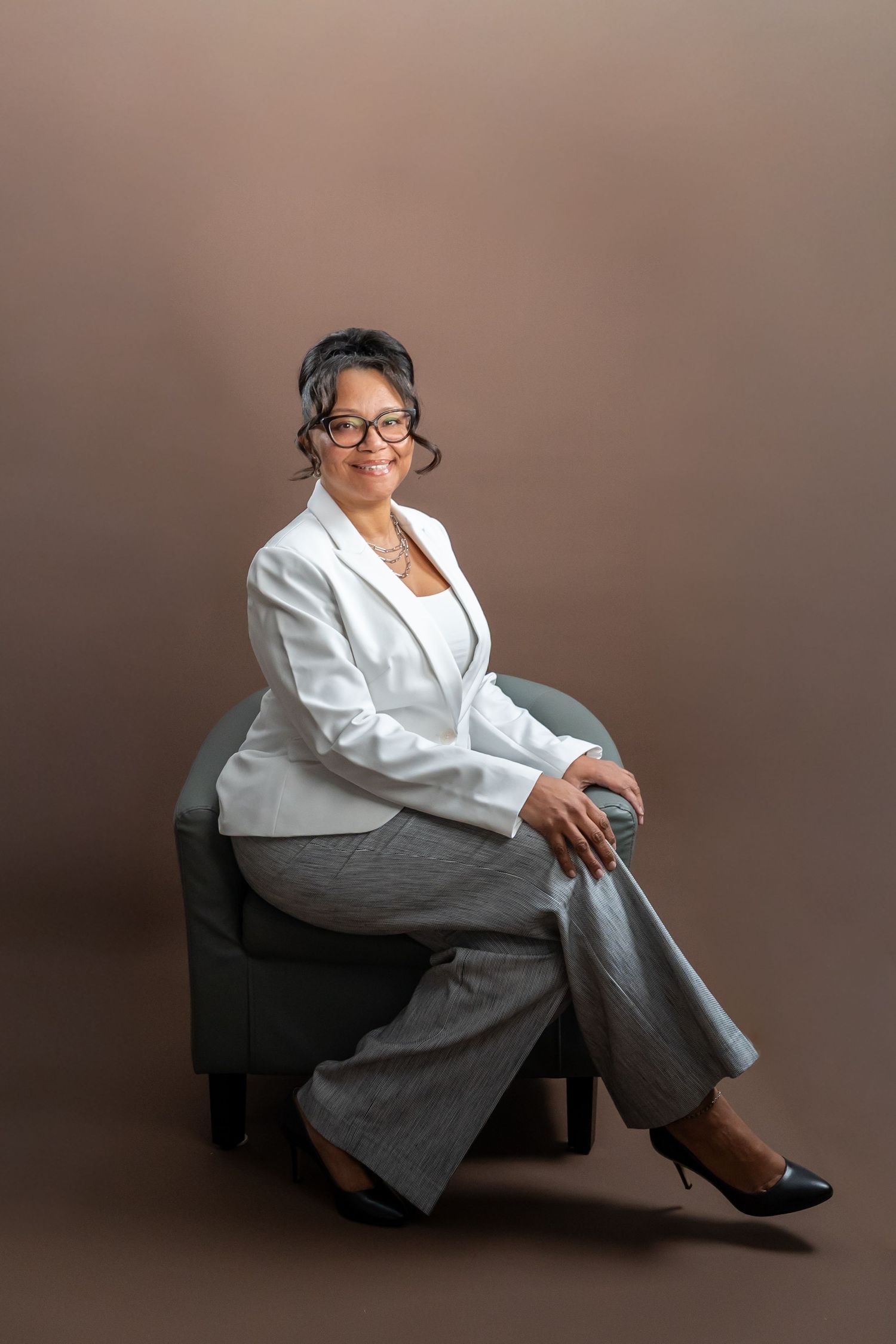 Professional woman in white blazer and gray pants sitting in chair against brown background.