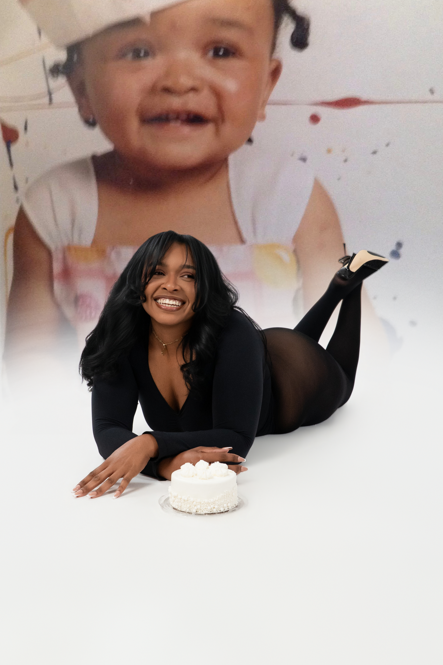 A smiling woman in a black outfit lies on a white floor beside a small cake, with a baby portrait backdrop behind her.