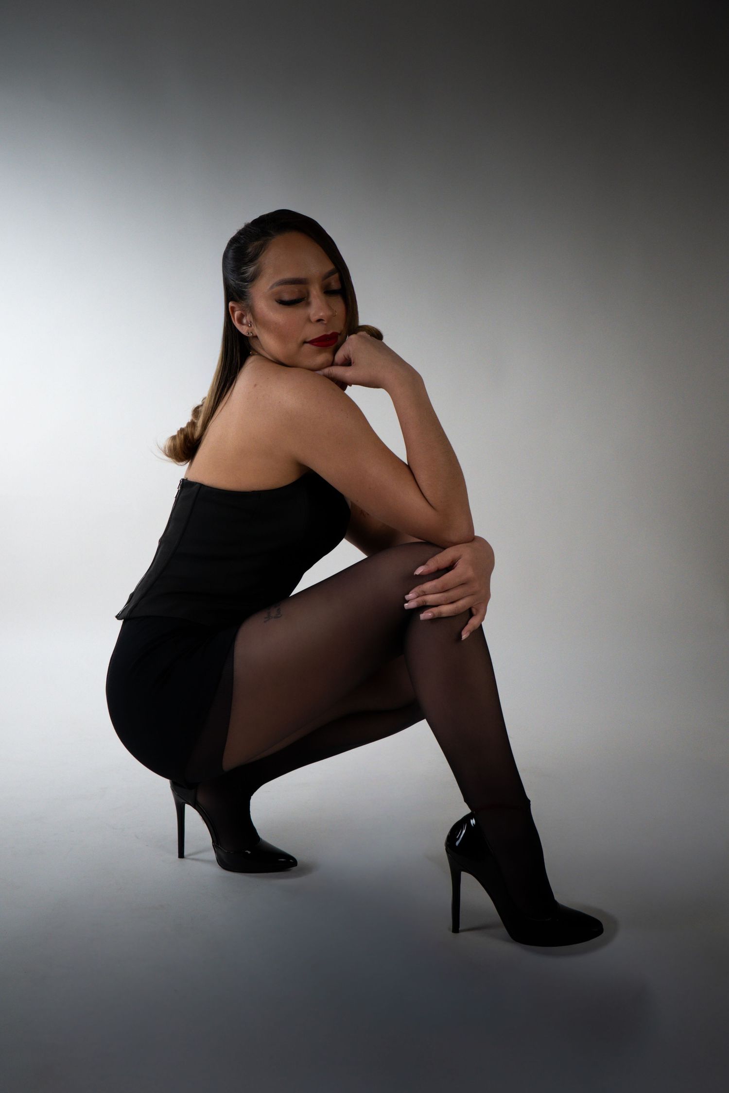 Woman in black strapless mini dress and heels posing gracefully in a crouching position on studio background.