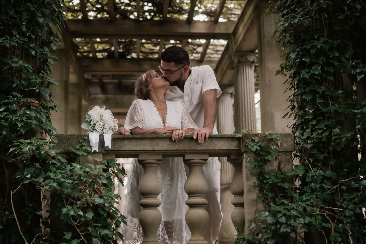 Couple shares a romantic kiss on a stone balcony surrounded by lush green ivy in a garden setting.
