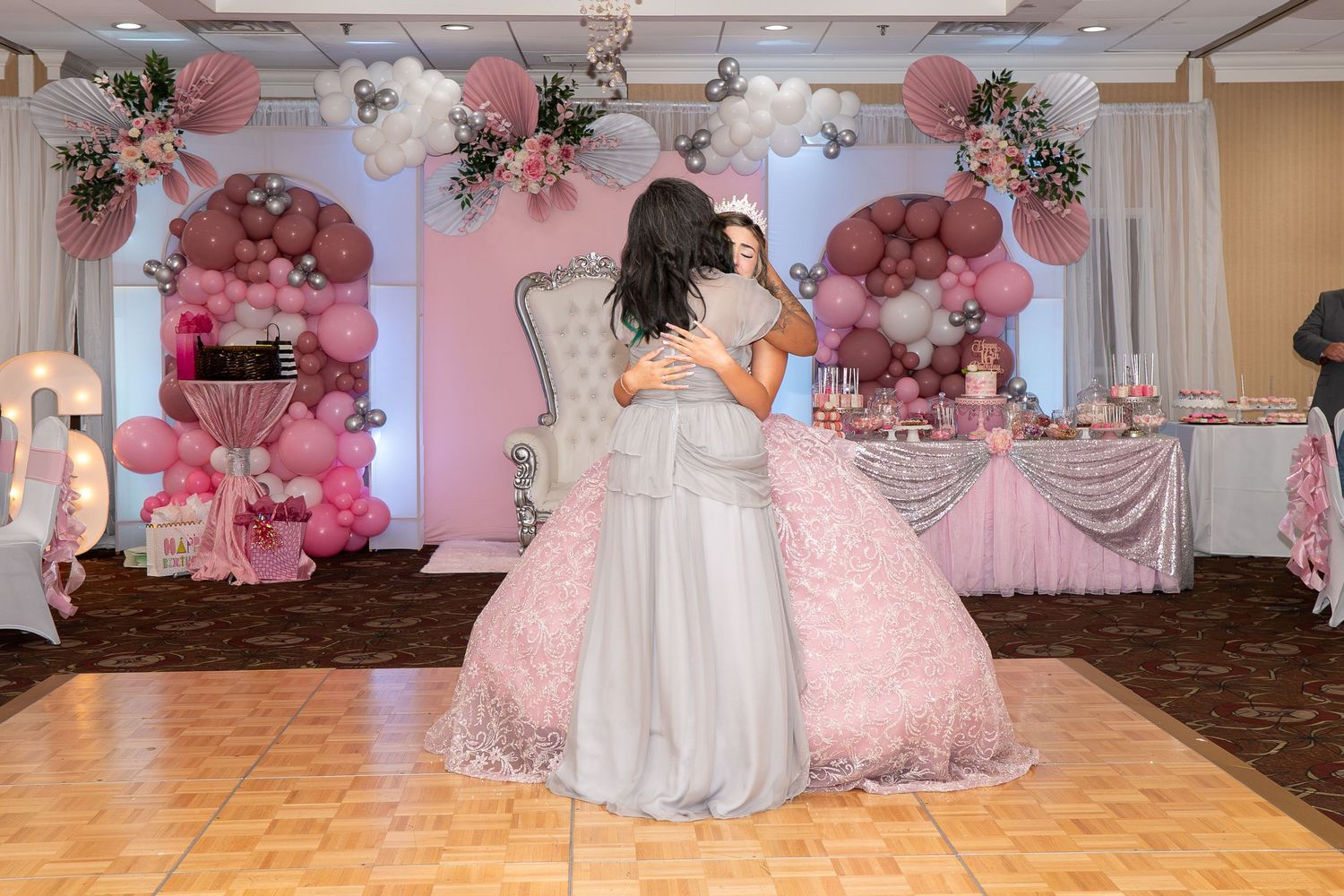 Two girls embrace on a dance floor at a pink and silver quinceañera celebration with balloon arch decor. Sweet 16!