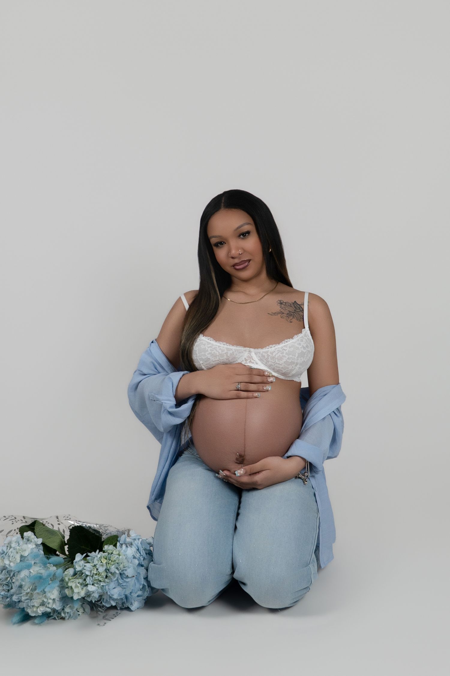 Pregnant woman in white lace bralette and jeans sits gracefully with blue hydrangeas, cradling her baby bump.