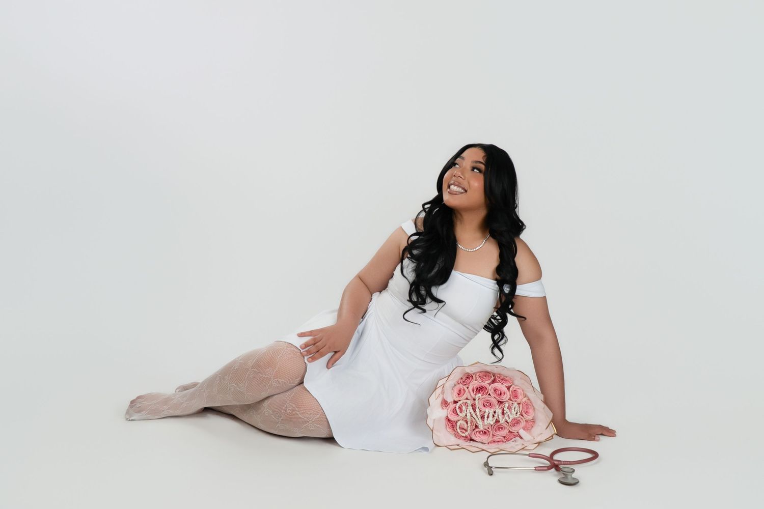 Woman in white dress sitting on floor with pink floral arrangement and scissors on white background.