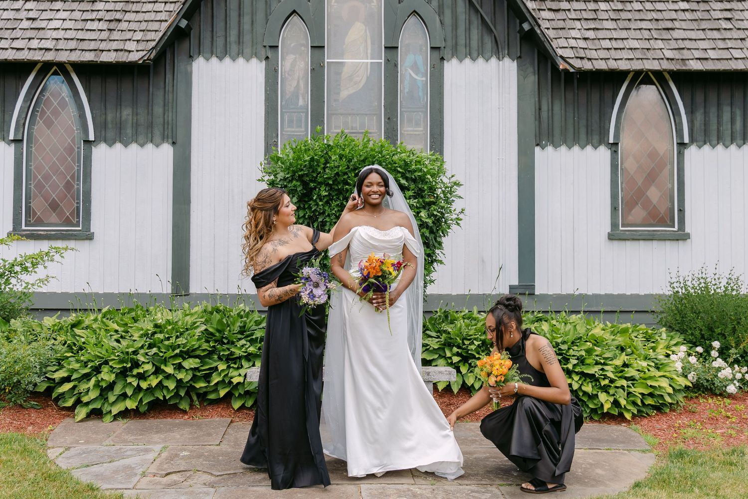 Bride in white gown with two bridesmaids in black dresses holding colorful bouquets outside a charming white chapel.