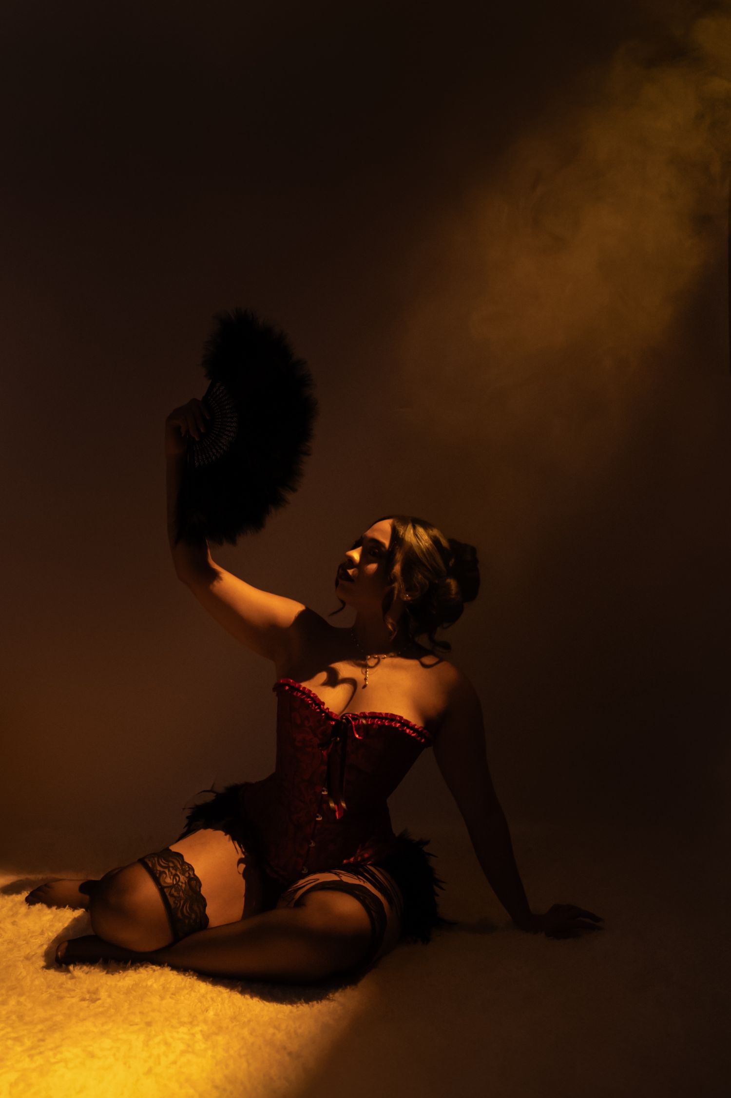 A woman in a red sequin corset and masquerade mask poses dramatically with a black feather fan in moody golden light.