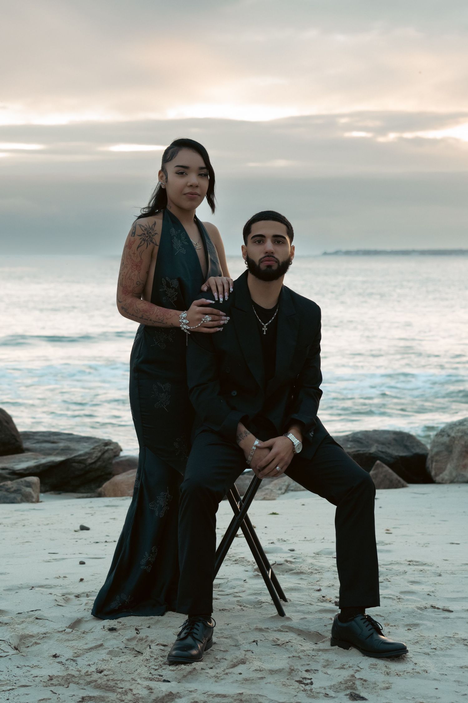 Elegant couple in black formal wear posing on beach at sunset.
