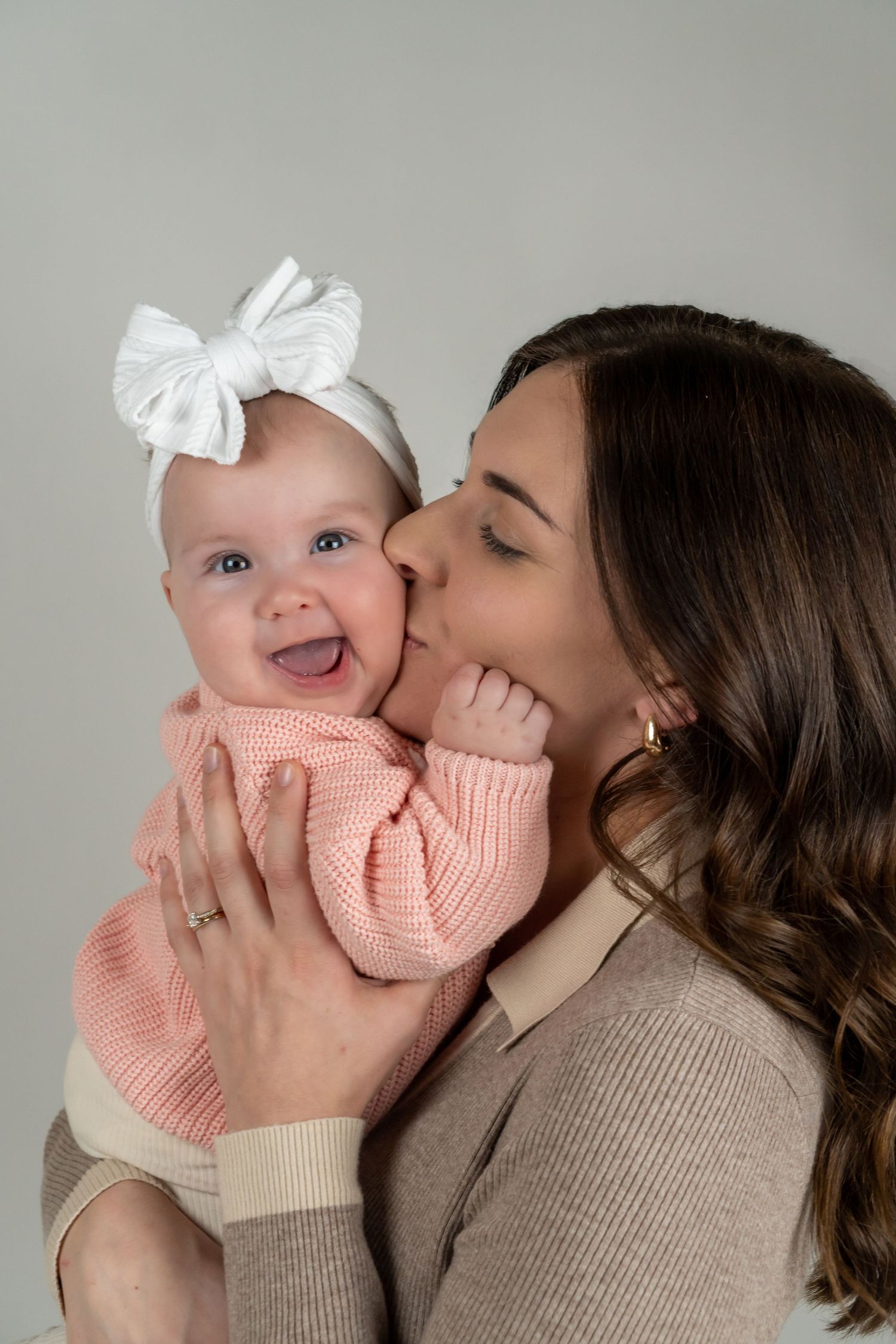 Mother kissing smiling baby girl wearing white bow headband and pink knit outfit.