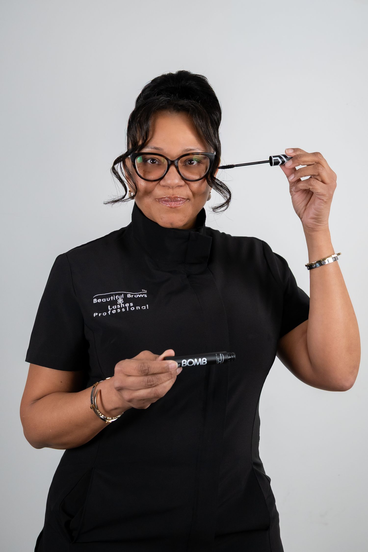 Female beauty professional in black uniform holding mascara wand and makeup tool in studio portrait.