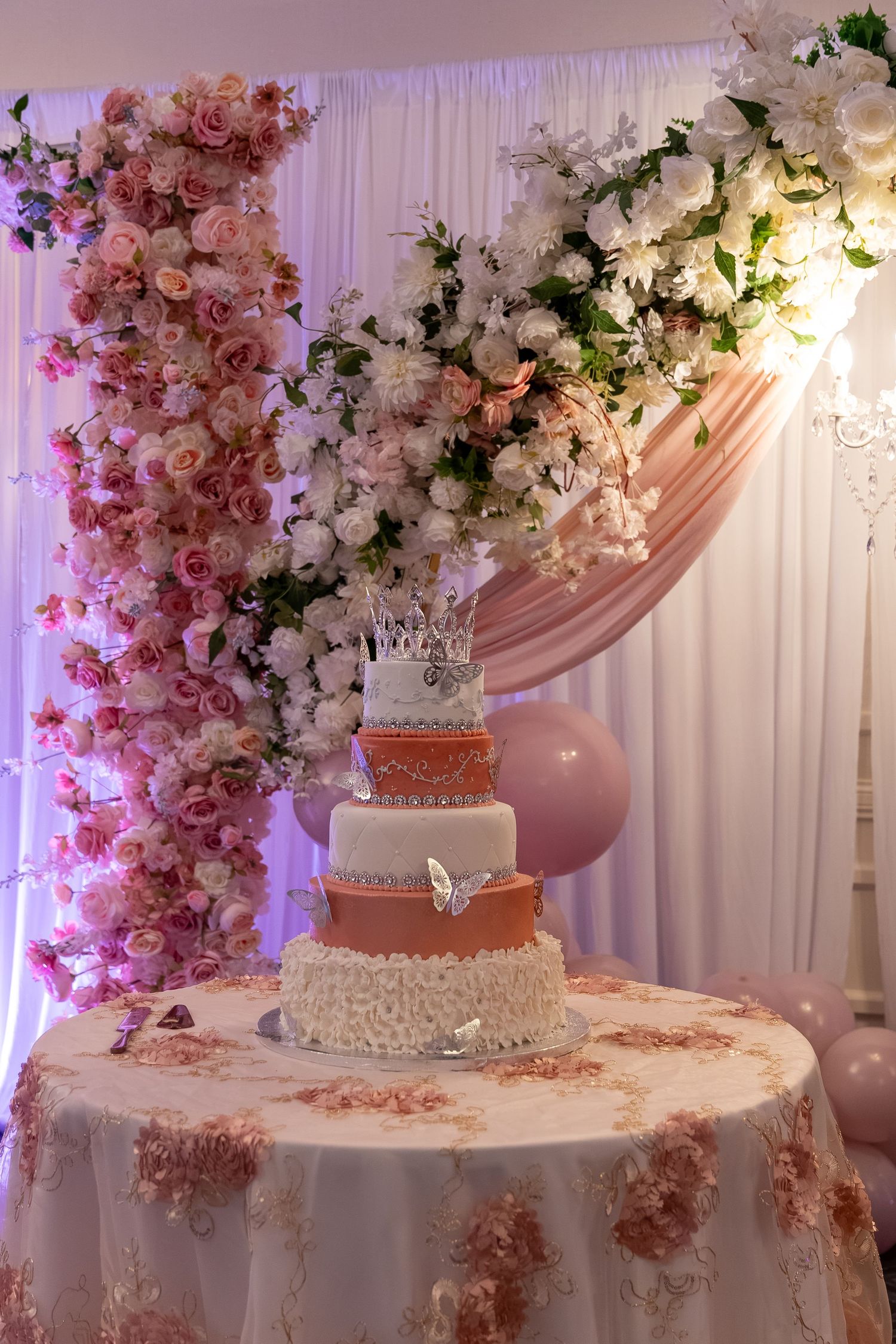 Elegant three-tier wedding cake with crown topper on floral table, surrounded by pink and white flower arch backdrop. Sweet 16/Quinceañera